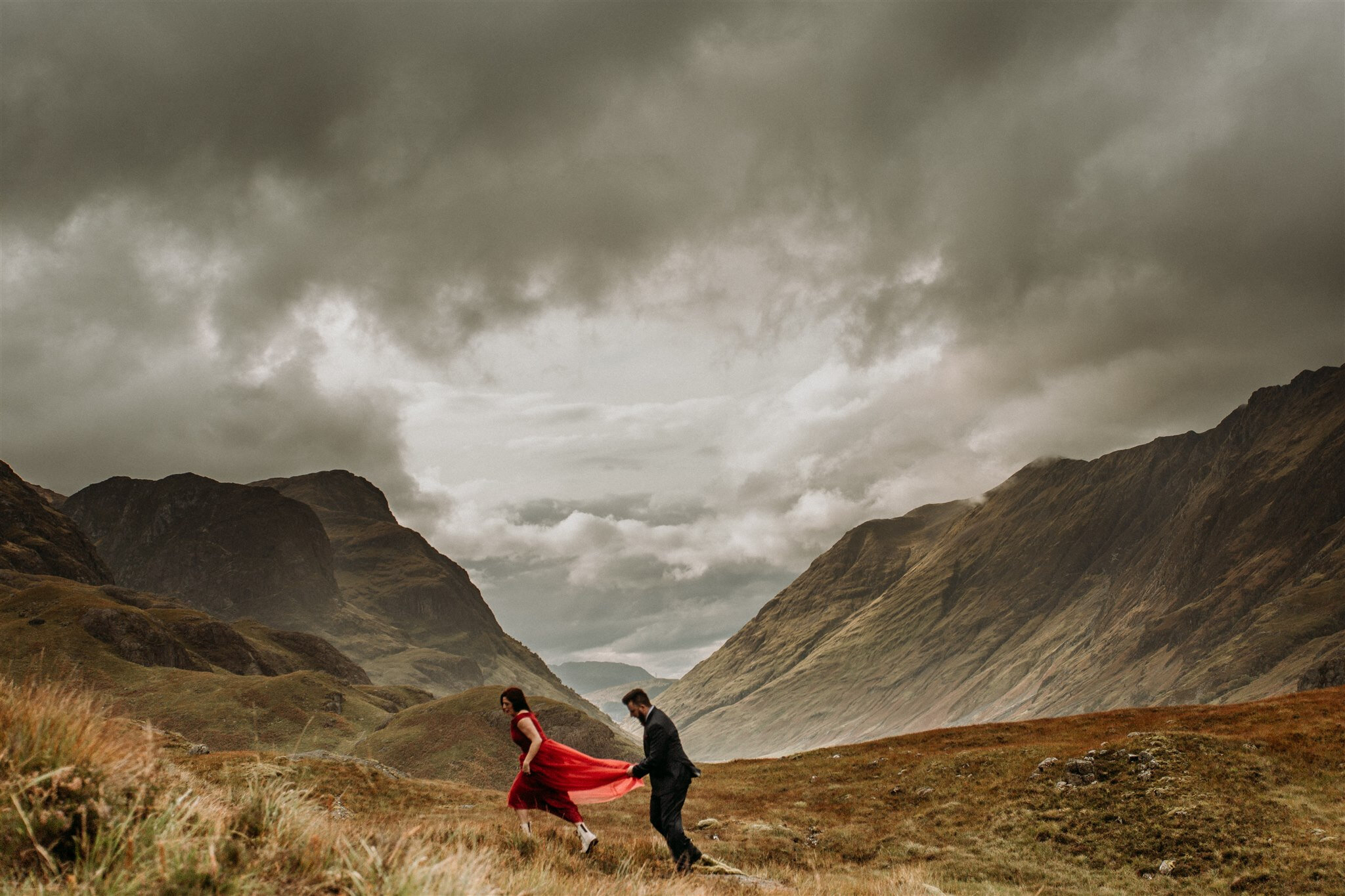 Glen Coe Scotland adventure elopement session. Bride in red dress and her groom walking hand in hand through a windy field in the Scottish Highlands | Adventure elopement photographer