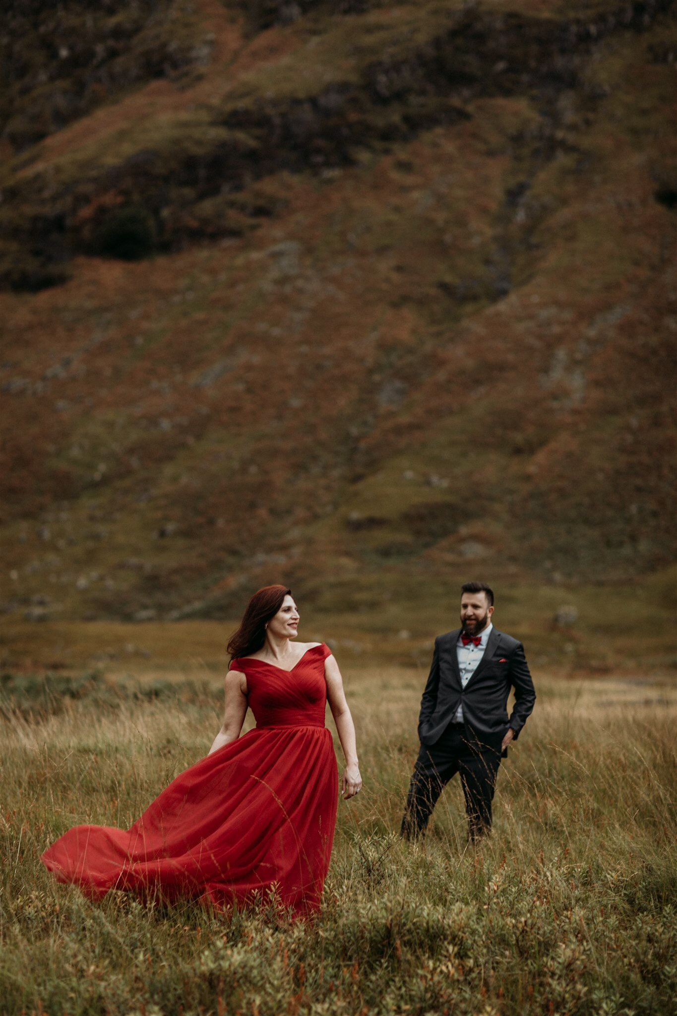 Glen Coe Scotland elopement adventure session. Bride in red dress blowing in the wind and her groom stand slightly apart in a field in the Scottish Highlands | Adventure elopement photographer