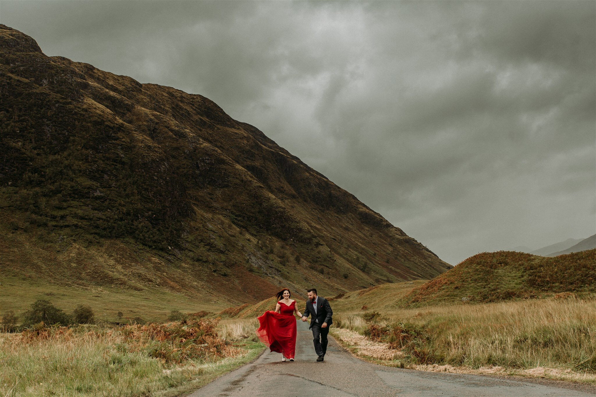 Glen Coe Scotland adventure elopement session. Bride in red dress and her groom walk hand in hand down a road in the windy Scottish Highlands | Adventure elopement photographer