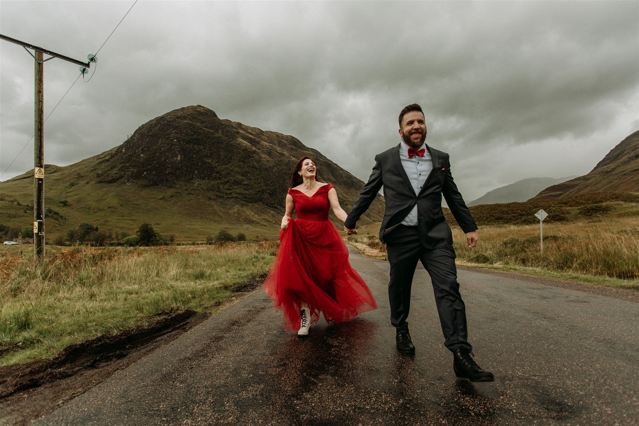 Glen Coe Scotland adventure elopement session. Bride in red dress and her groom walk hand in hand down a road in the windy Scottish Highlands | Adventure elopement photographer