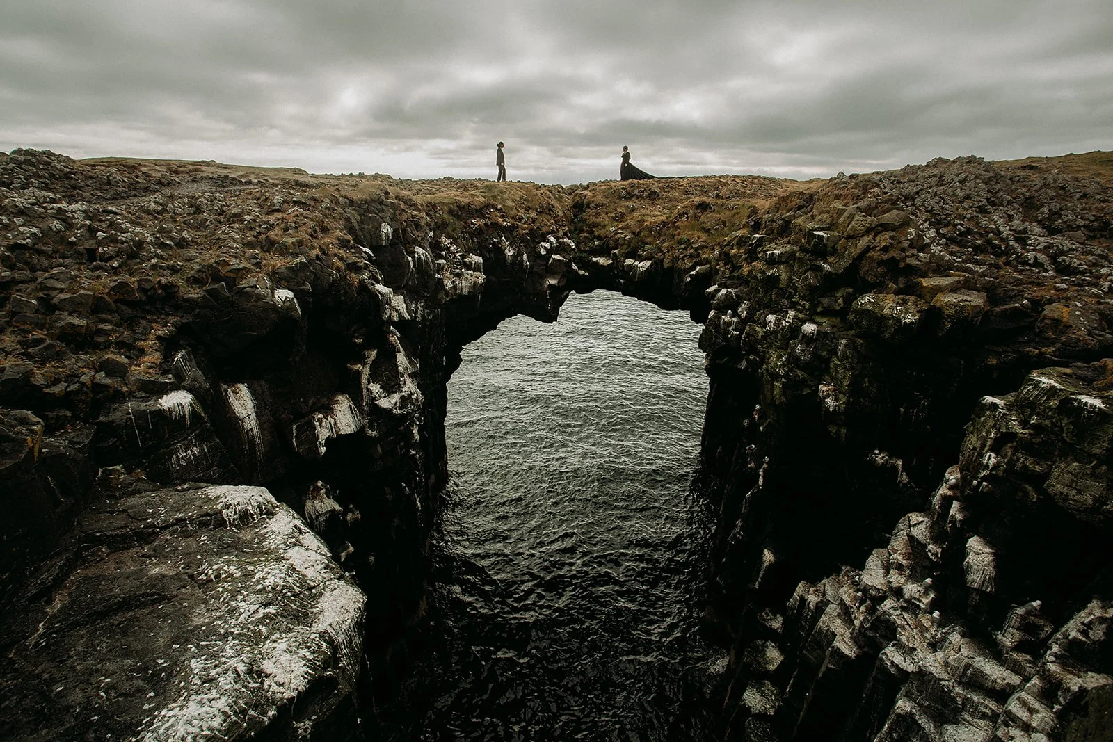 Alternative Iceland elopement couple wearing black wedding dress and purple hair — couple portraits sea cliffs Snæfellsnes rock bridge