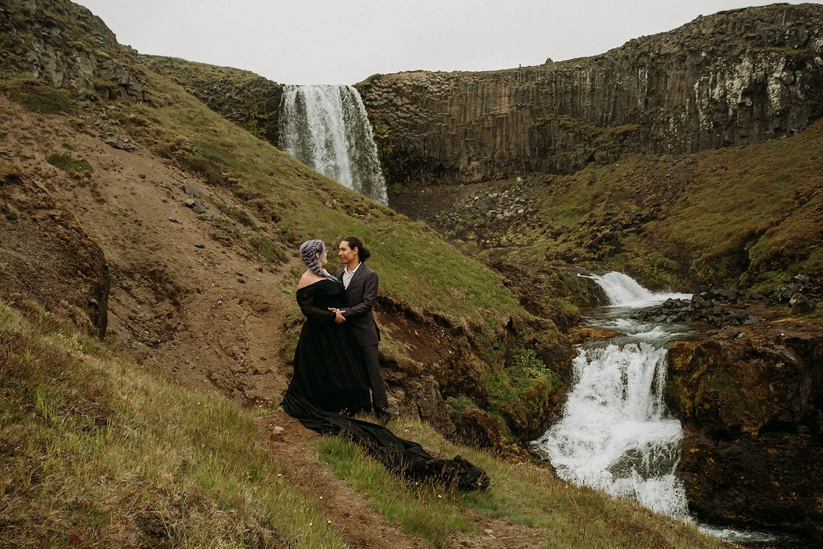Alternative Iceland elopement couple wearing black wedding dress and purple hair — couple portraits at waterfall 