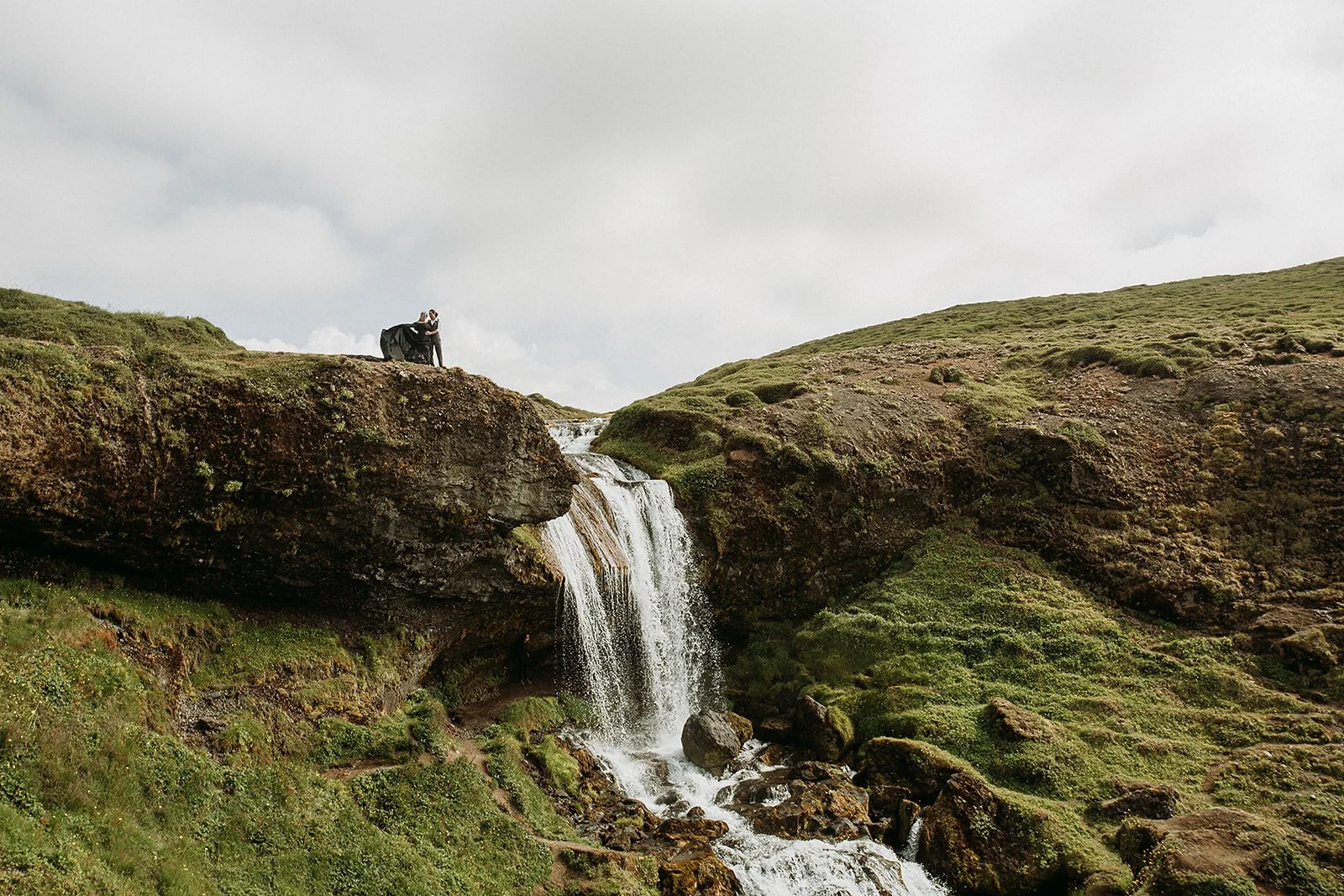 Alternative Iceland elopement couple wearing black wedding dress and purple hair — couple portraits at waterfall 
