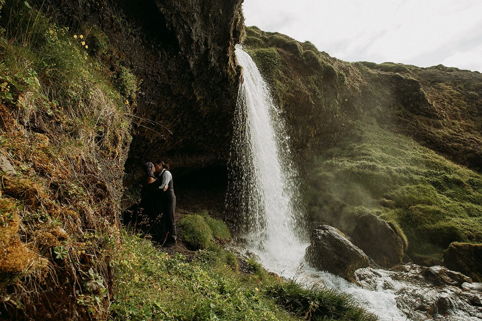 Alternative Iceland elopement couple wearing black wedding dress and purple hair — couple portraits at waterfall 