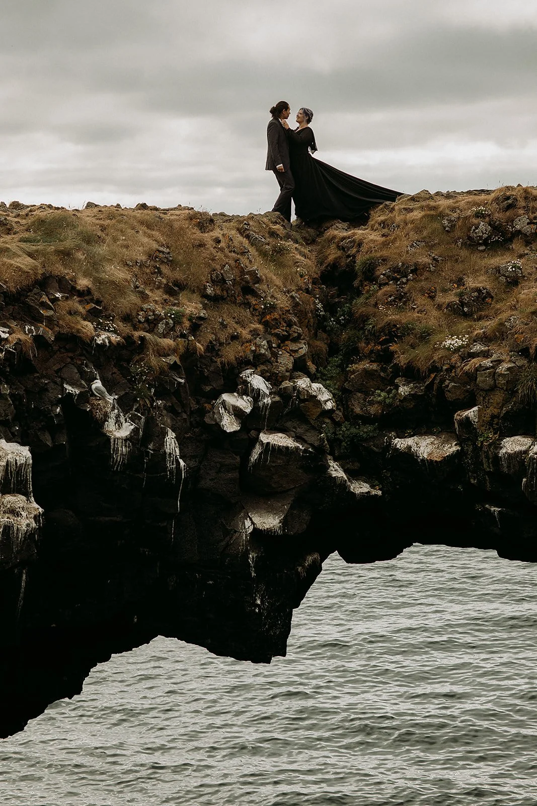 Alternative Iceland elopement couple wearing black wedding dress and purple hair — couple portraits sea cliffs Snæfellsnes rock bridge