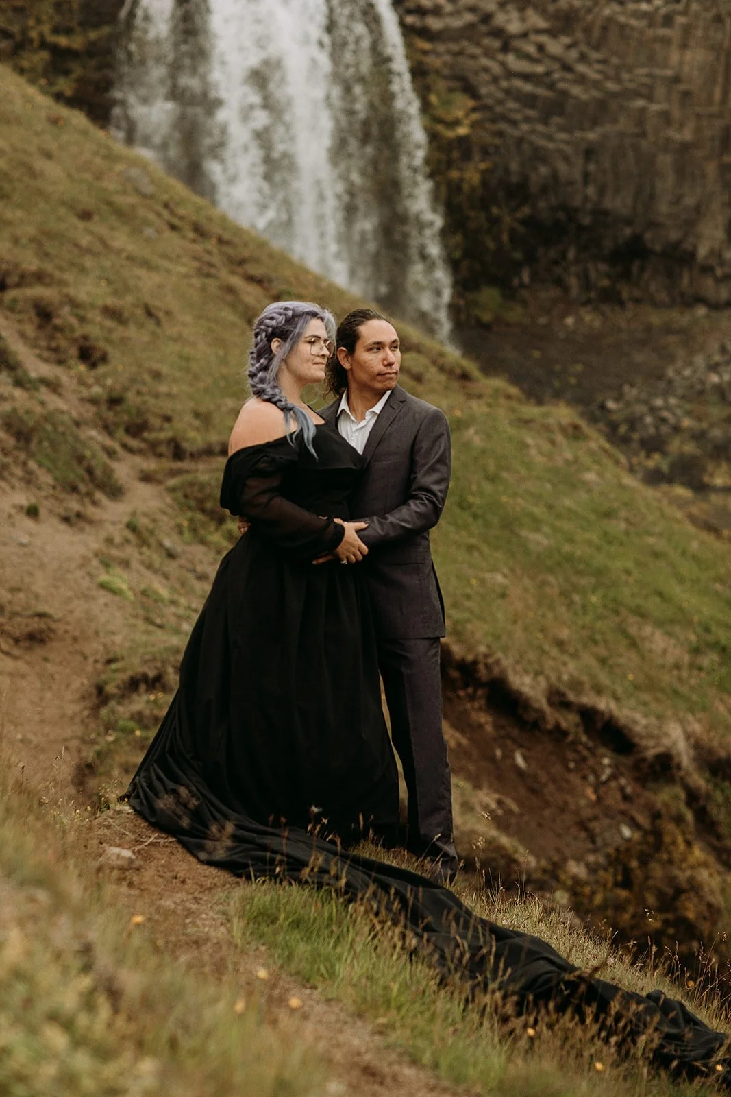 Alternative Iceland elopement couple wearing black wedding dress and purple hair — couple portraits at waterfall