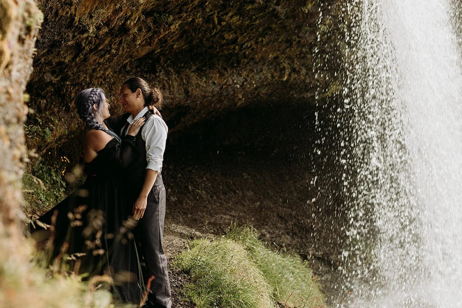 Alternative Iceland elopement couple wearing black wedding dress and purple hair — couple portraits at waterfall