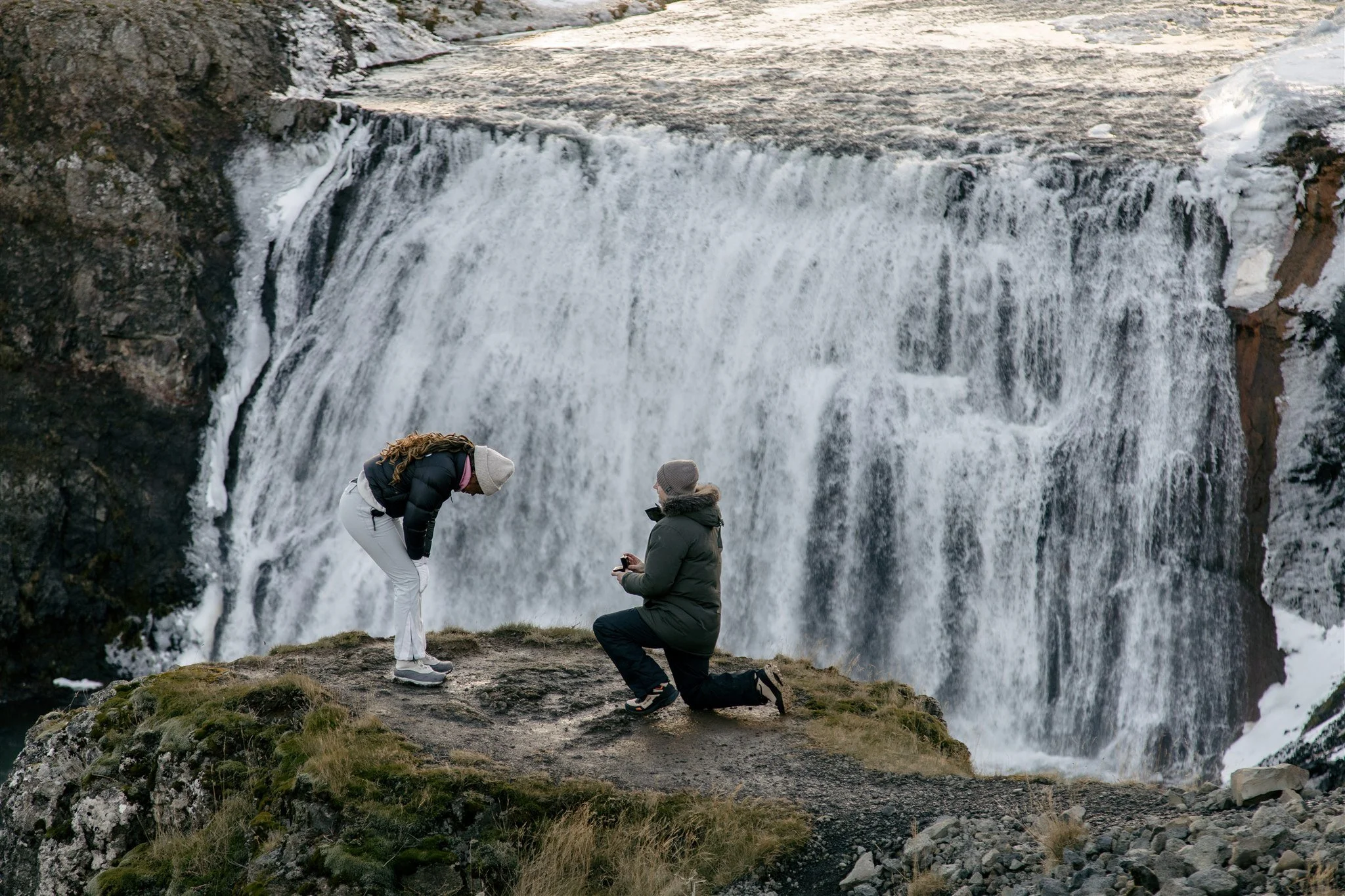 Iceland Marriage Proposal Image: A couple gets engaged in Iceland on vacation with a stunning backdrop with one partner on one knee and the partner being proposed to is so happy bending over in disbelief .