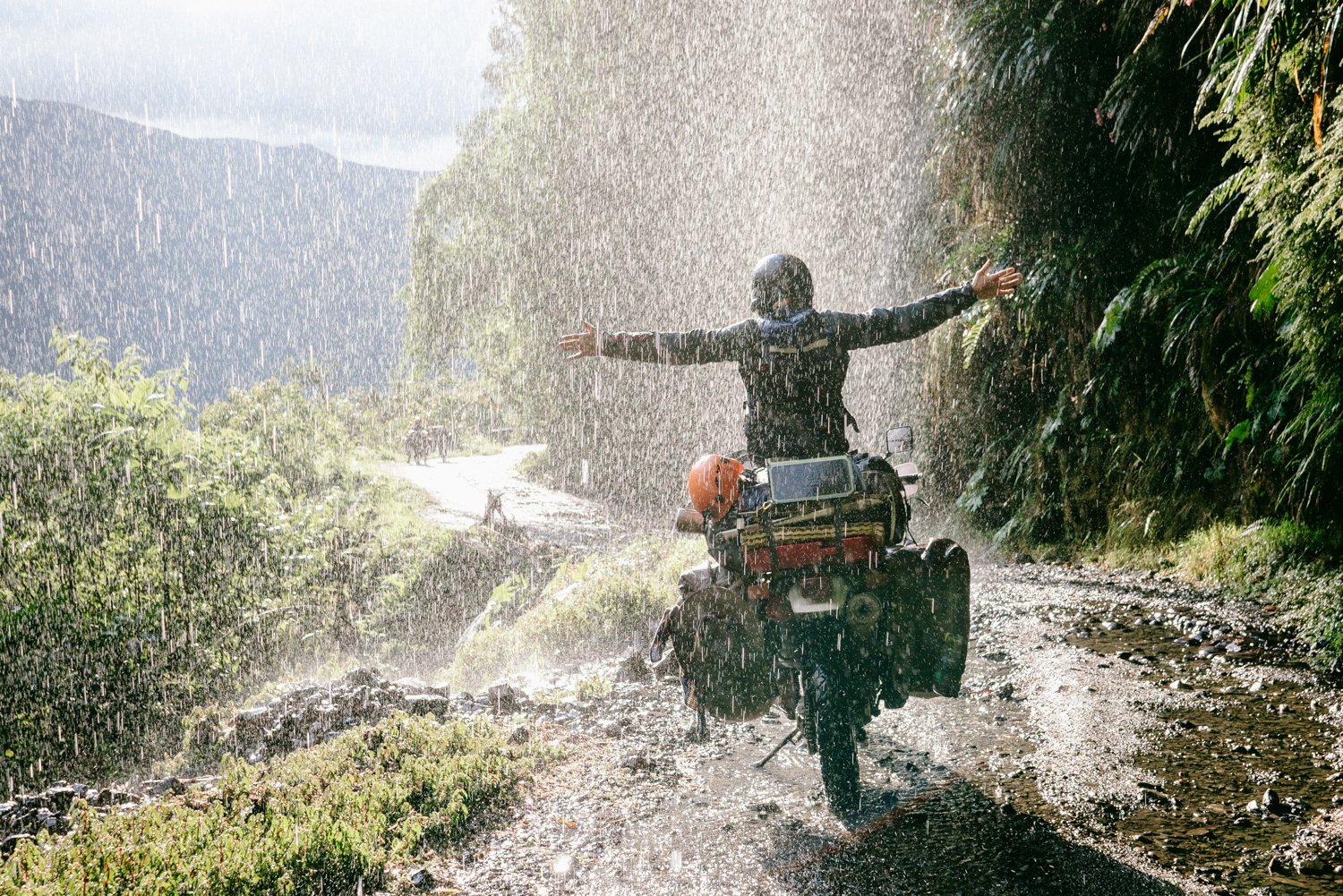  Motorcycle with rider holding hands out straight to sides on a lush mountain trail during a sunshower. 