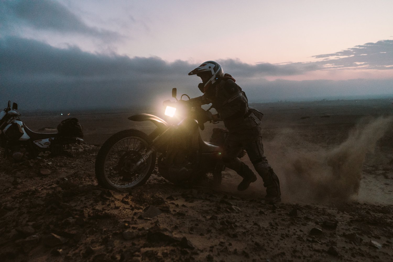  Motorcycle at dusk stuck in mud as the rider stands beside it trying to get it out. 