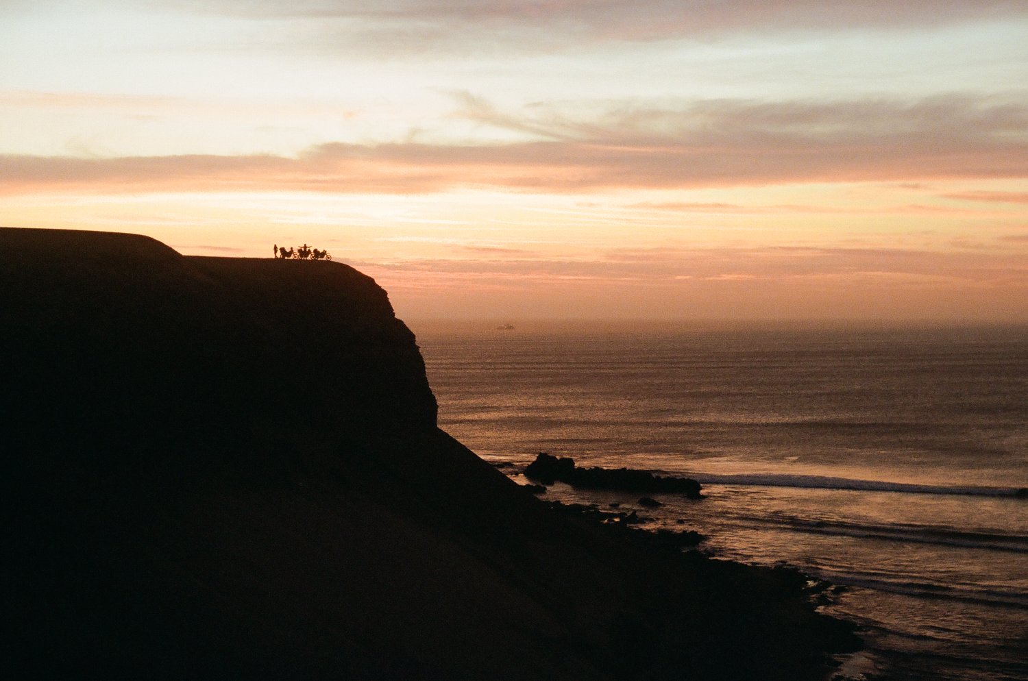  Sunset with three motorcycles and riders on a rock cliff high above the ocean.     