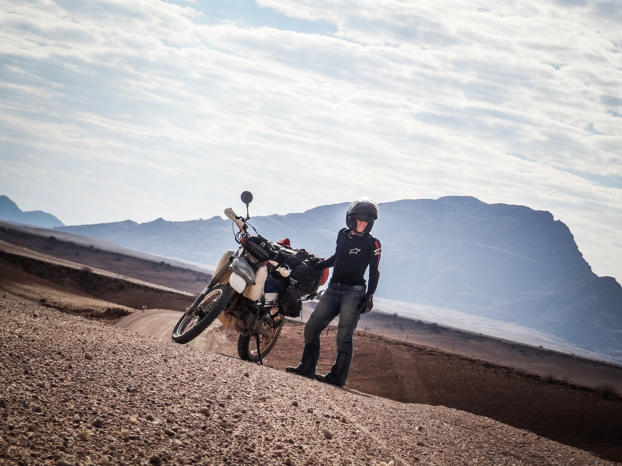  Woman rider standing beside motorcycle in a desert like area with mountains in background. 