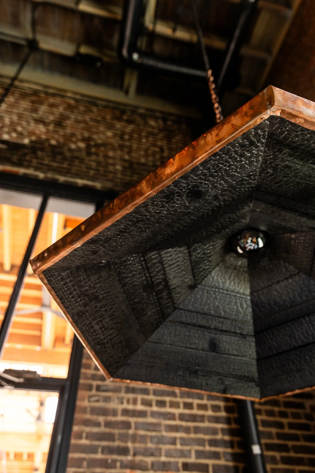 Close-up of a rectangular hanging table with a metallic frame, seen from below, with exposed brick wall and ceiling in the background.
