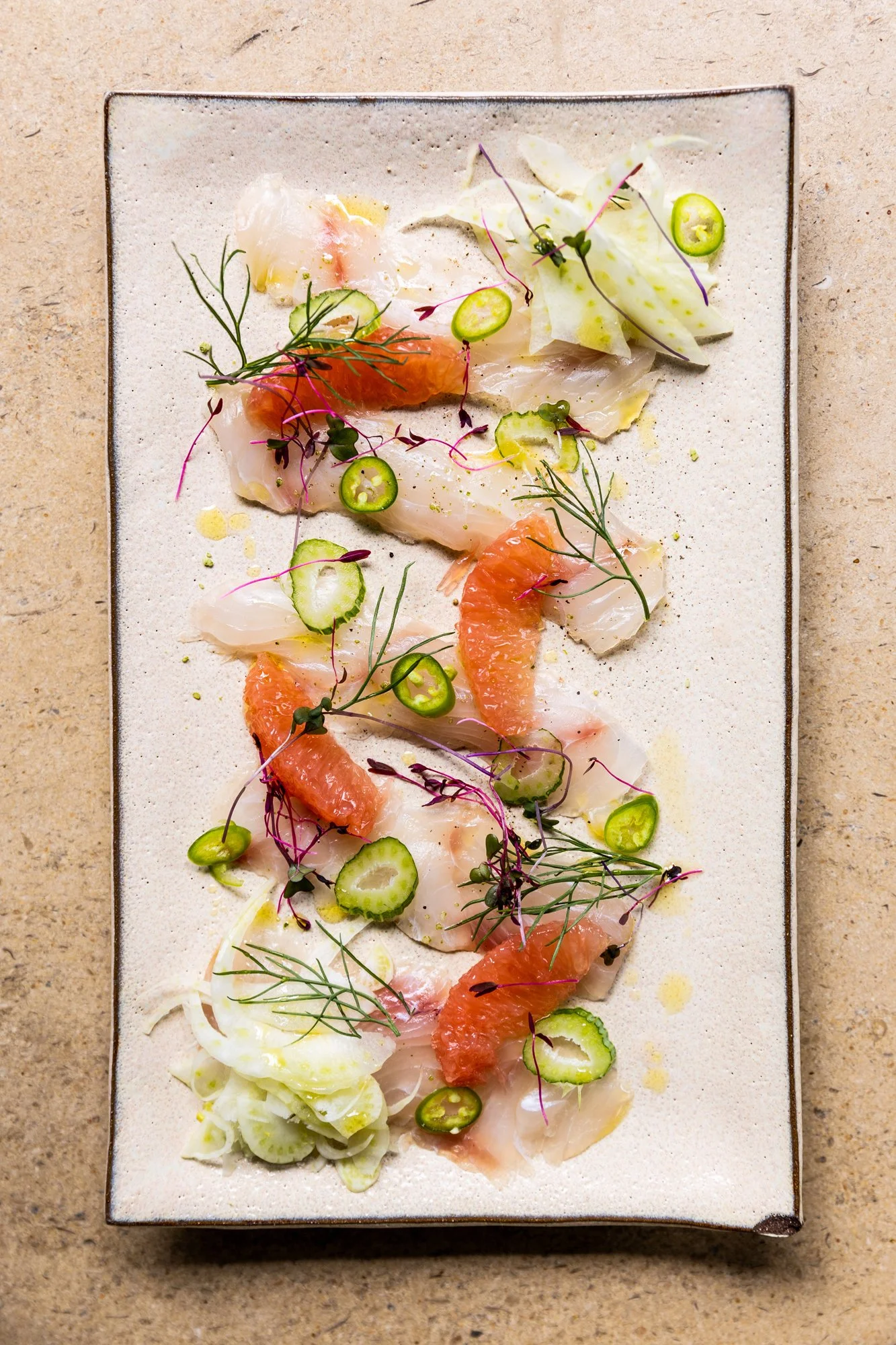 Seafood dish featuring slices of fish, citrus segments, cucumber slices, microgreens, and thinly sliced onions, presented on a rectangular plate. Hot and Hot Fish Club Restaurant in Birmingham, Alabama.