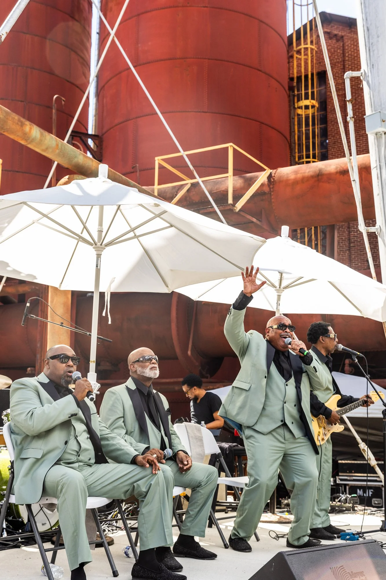A blues band performing outdoors with three male singers in matching light green suits, one singing into a microphone with his hand raised, and a guitarist playing beside them. Outdoors with large white umbrellas and overcast sky.