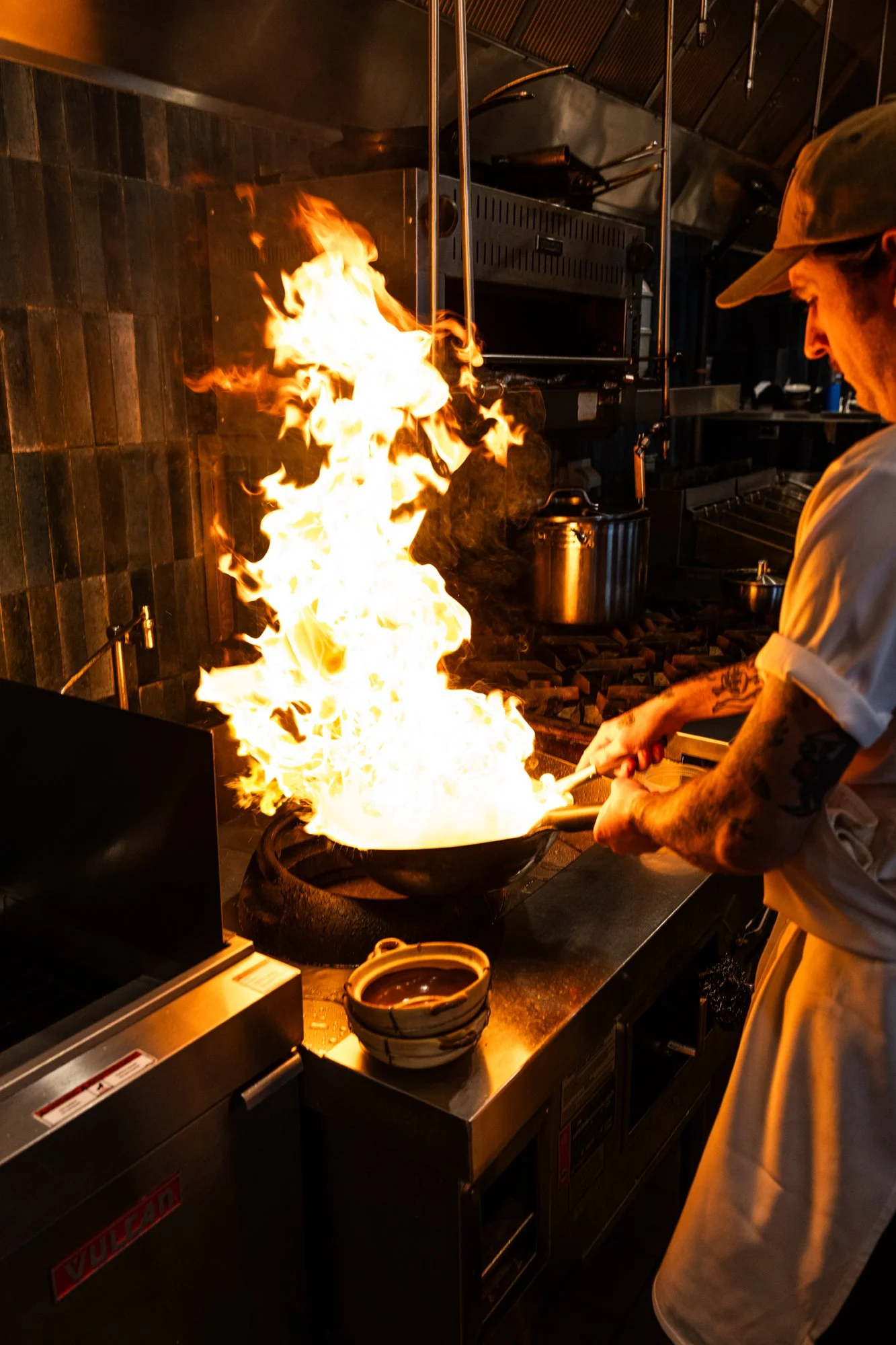 A chef in a restaurant kitchen is cooking with a large flame in a frying pan, creating a fire display as part of a culinary technique.