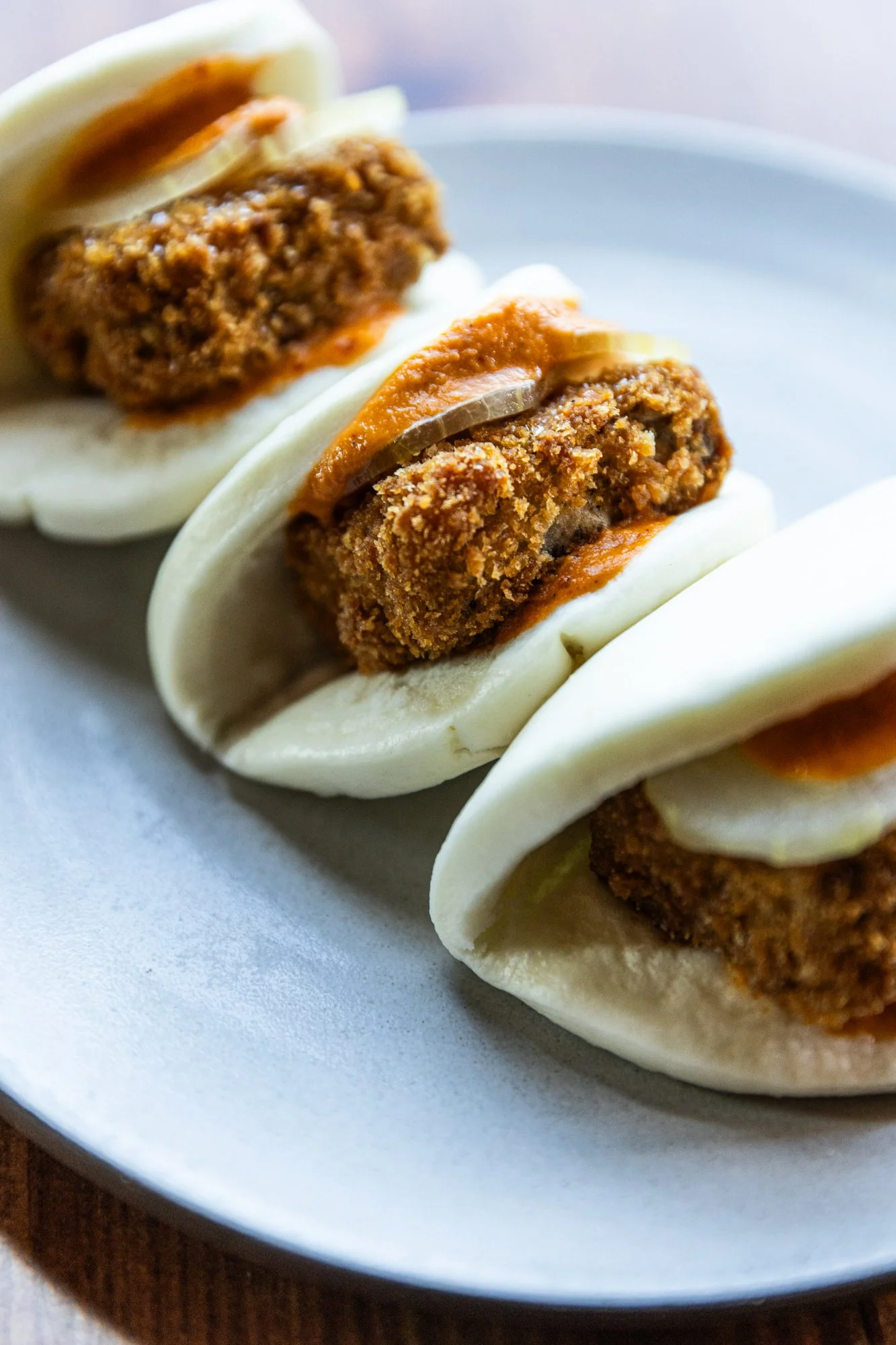 Close-up of three steamed pork buns with crispy fried pork, pickles, and chili sauce, served on a white plate.