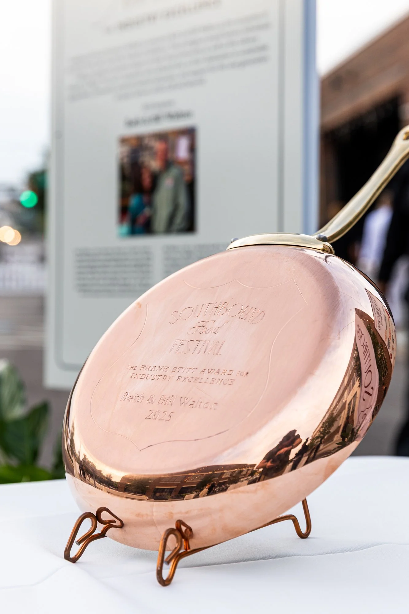Copper-colored anvil-shaped trophy with engravings, displayed on a table, with an informational poster in the background at an outdoor event. Southbound Food Festival in Birmingham, Alabama.