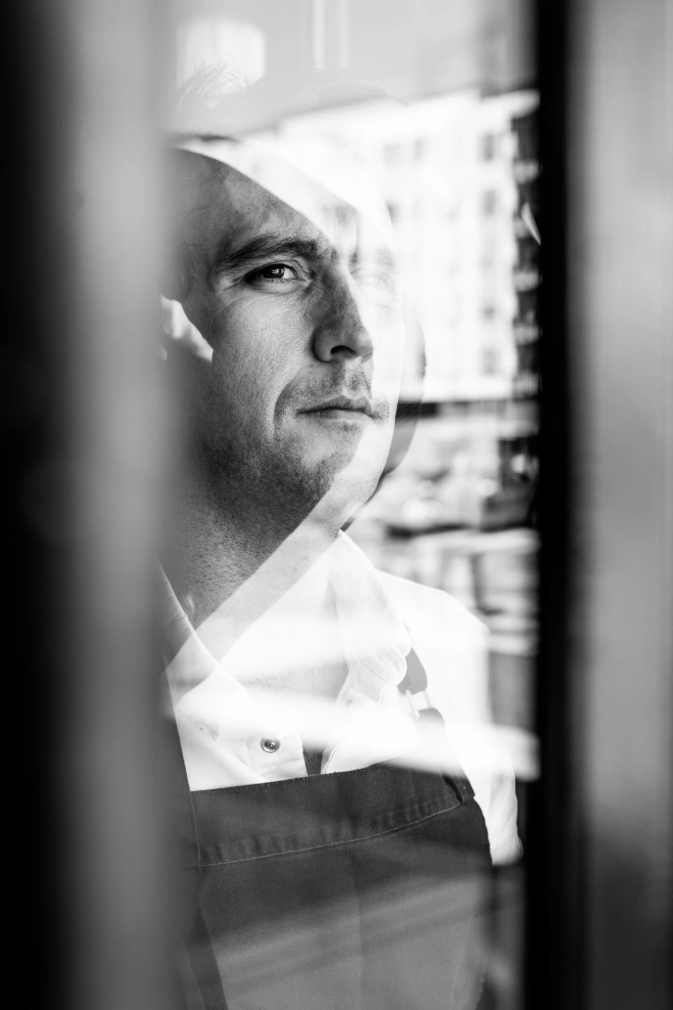 Black and white photo of a man looking through a window, wearing an apron, with a cityscape in the background.