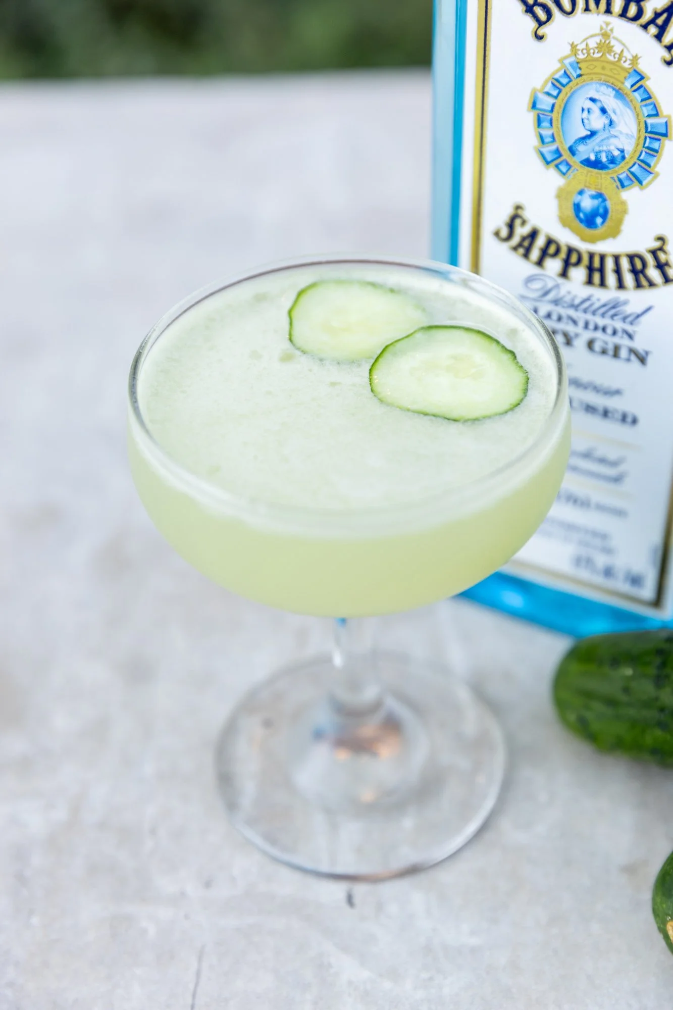 A pale green cocktail with two cucumber slices garnishing the top, in a coupe glass, with a bottle of Sapphire gin and fresh cucumbers in the background. Hot and Hot Fish Club Restaurant in Birmingham, Alabama.