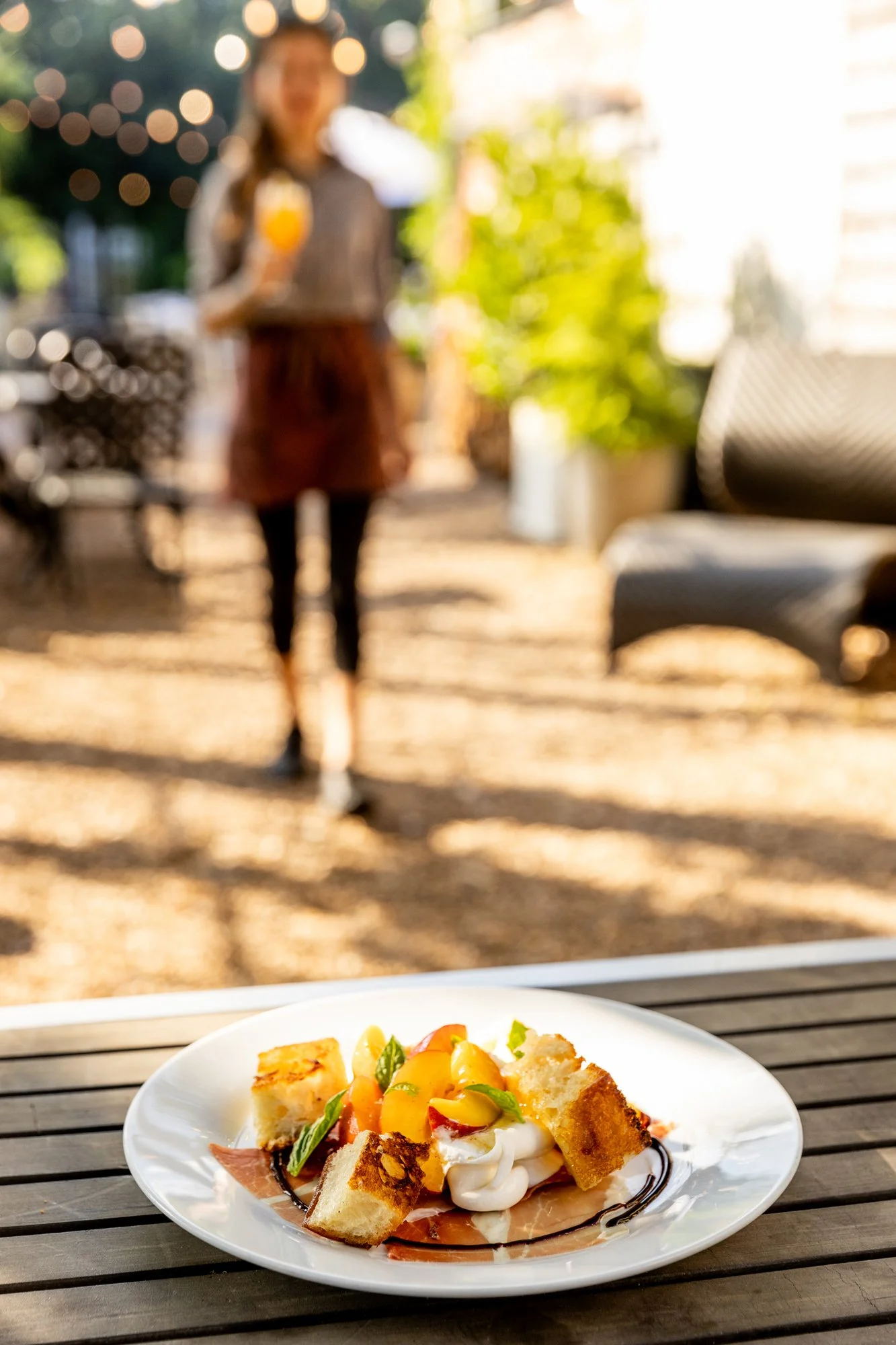 A white plate with a dessert that includes bread pieces, peaches, whipped cream, basil, and chocolate drizzle, on a wooden table outdoors, with a blurred woman holding a drink in the background. Ovenbird Restaurant in Birmingham, Alabama.