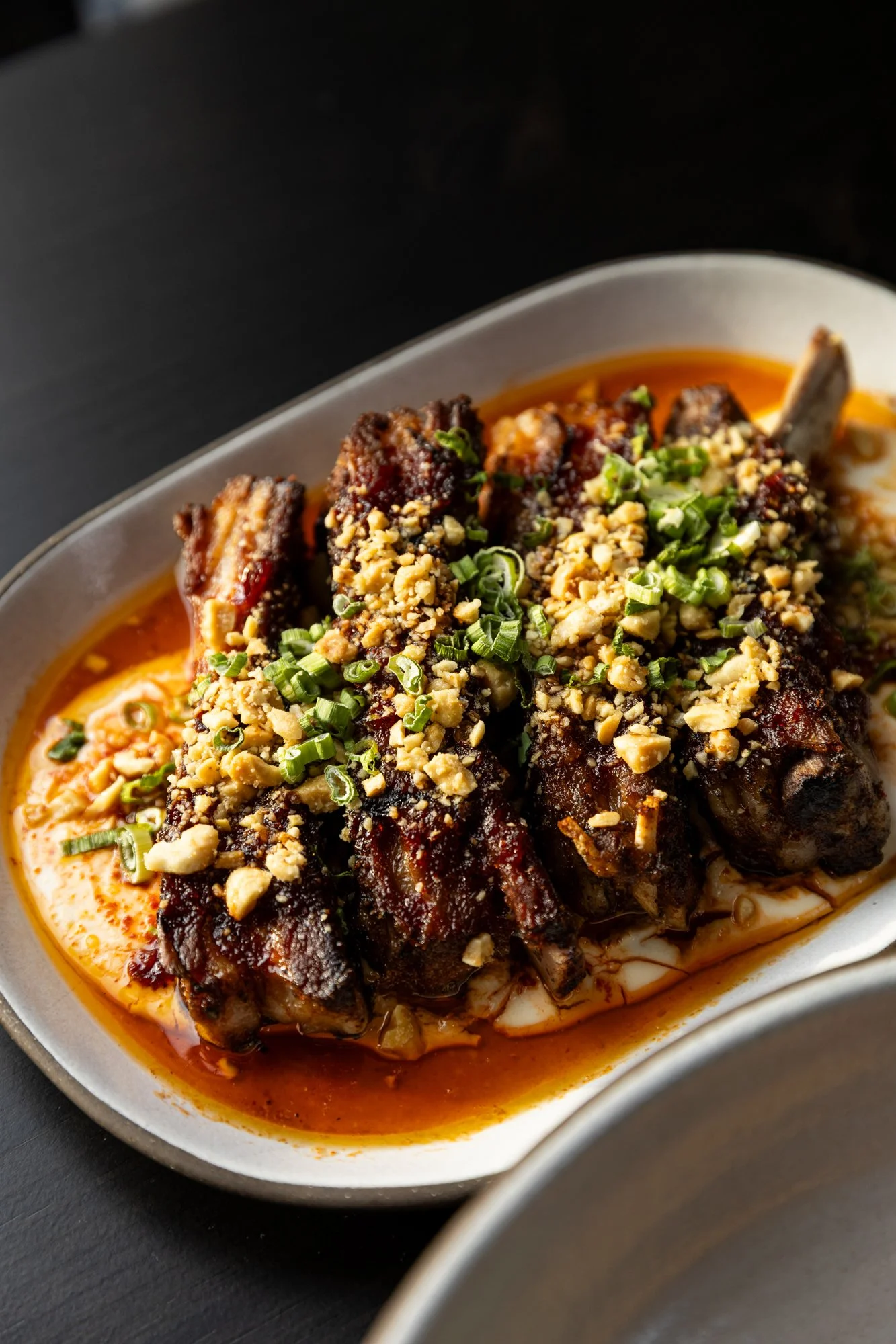 Close-up of grilled ribs topped with crushed nuts and chopped green onions, served in a creamy orange-colored sauce in a white oval dish. Birmingham, Alabama.