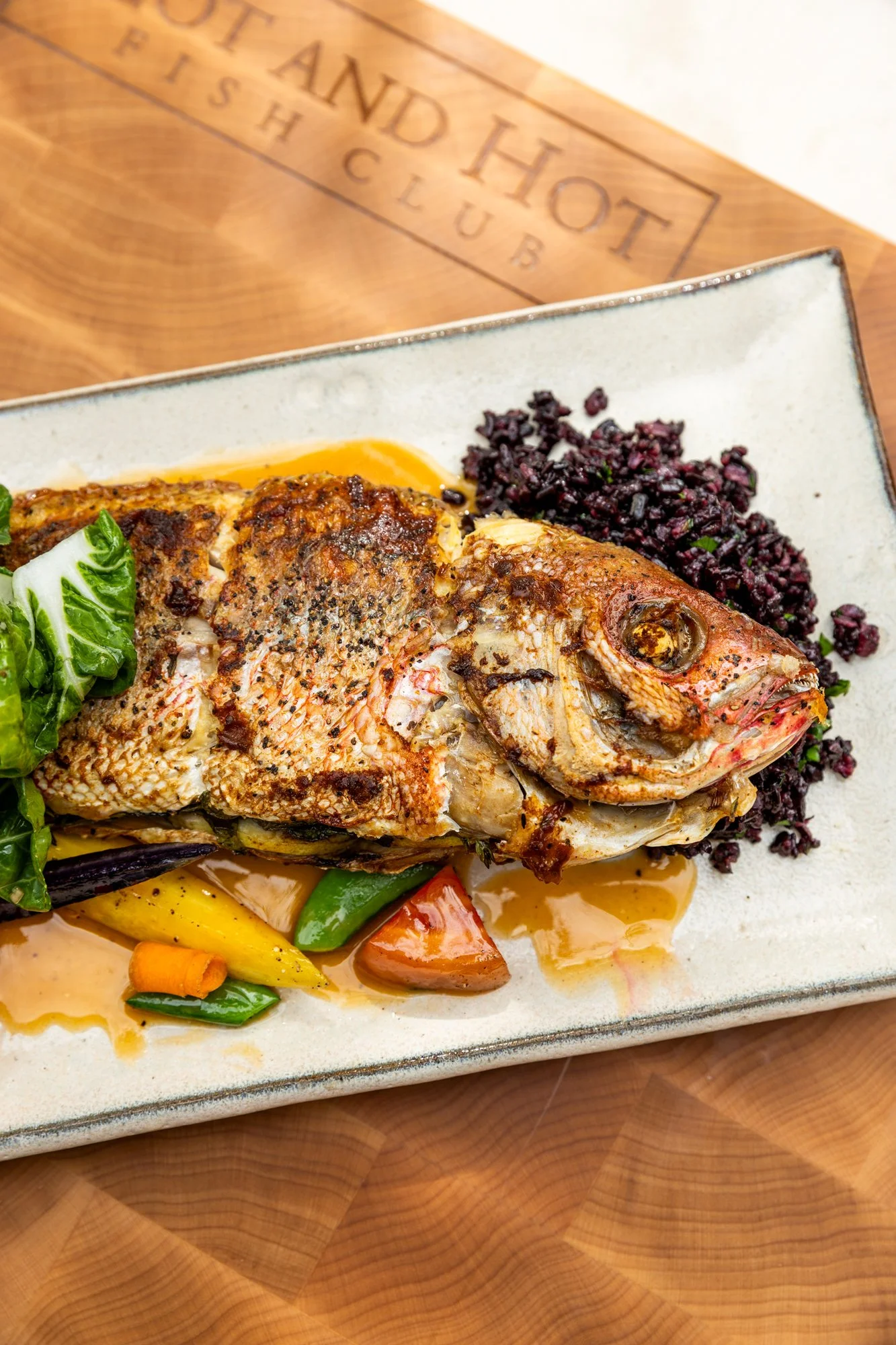 Whole cooked fish served on a plate with cooked vegetables, leafy greens, and purple rice, on a wooden table. Hot and Hot Fish Club Restaurant in Birmingham, Alabama.