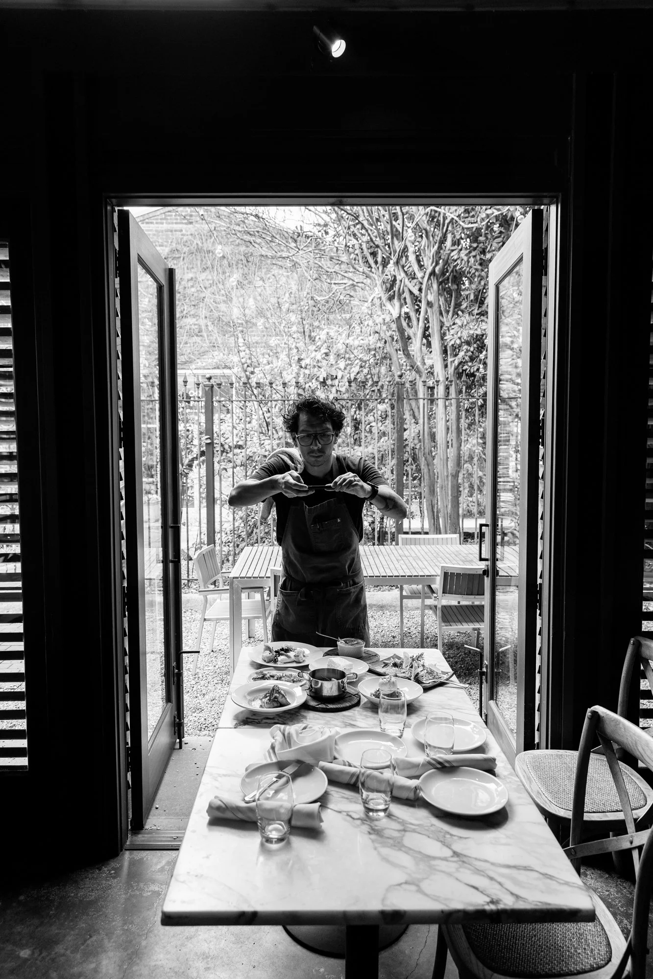Black and white photo of a man arranging a dinner table outdoors, viewed from inside through open glass doors. Ovenbird Restaurant in Birmingham, Alabama.