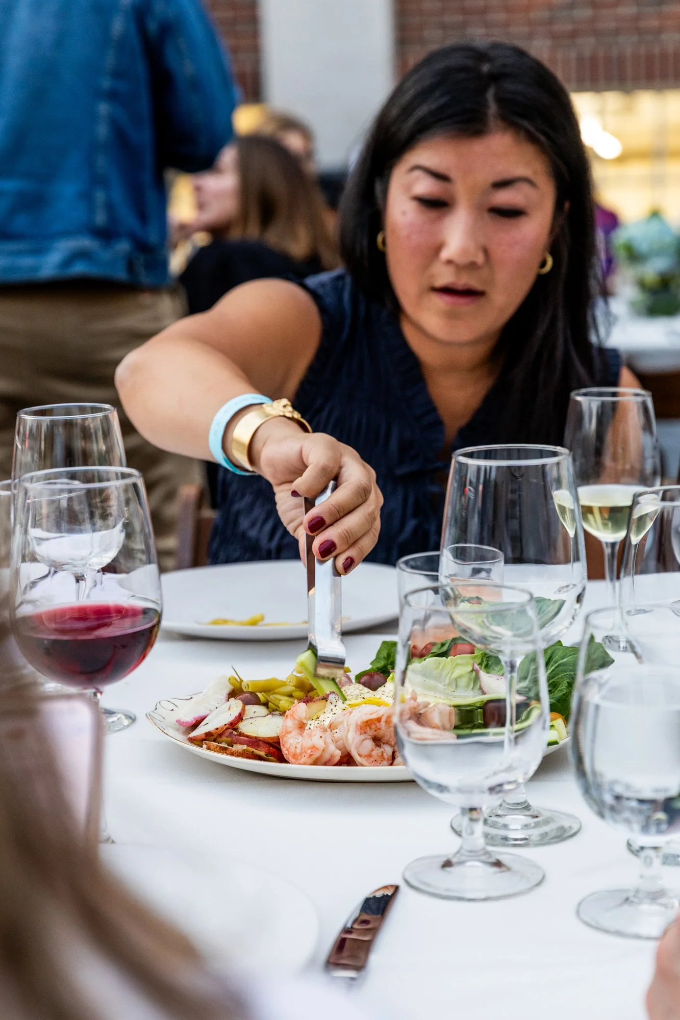 A woman in a dark blue dress is sitting at a table, serving herself food from a plate that has shrimp, vegetables, and other salad items. There are several glasses of wine and water on the table, along with a white tablecloth and a fork.