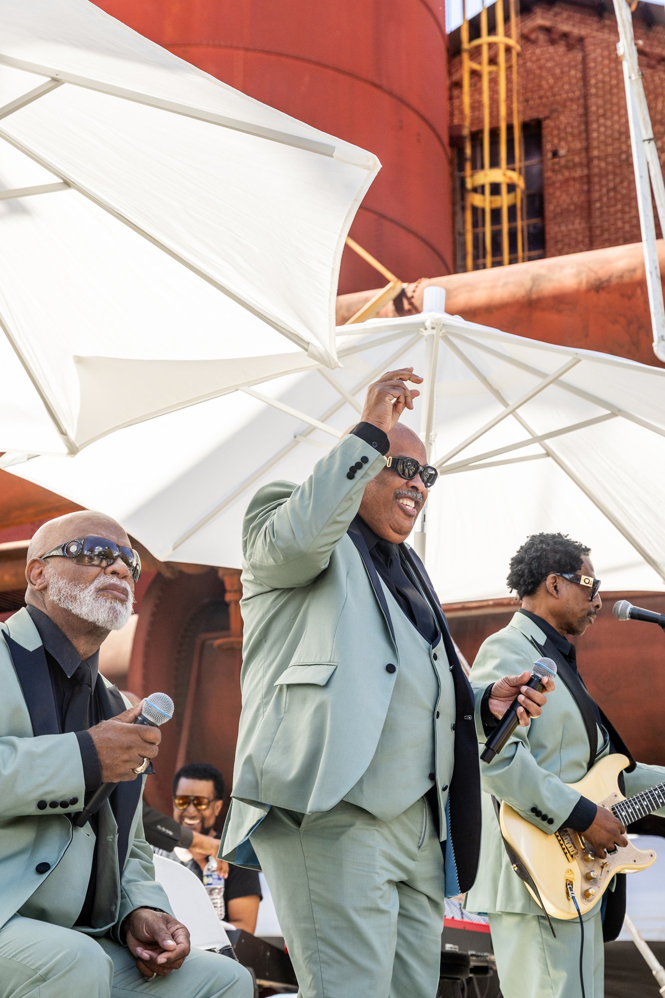 Music band performing outdoors, with members dressed in light-colored suits, wearing sunglasses, under large white umbrellas, with a reddish-brown building in the background. Southbound Food Festival in Birmingham, Alabama.