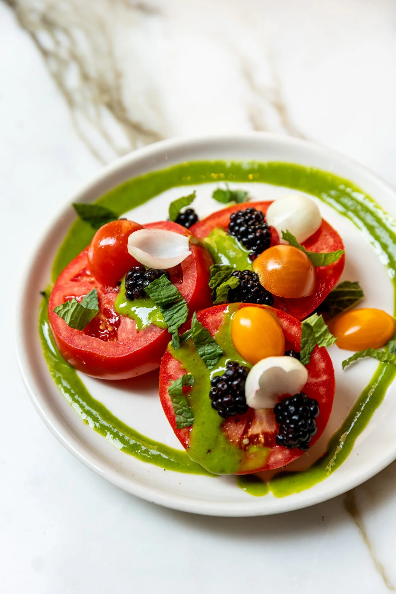 Close-up of a salad with tomato slices, yellow cherry tomatoes, blackberries, fresh herbs, and green dressing on a white plate. Ovenbird Restaurant in Birmingham, Alabama.