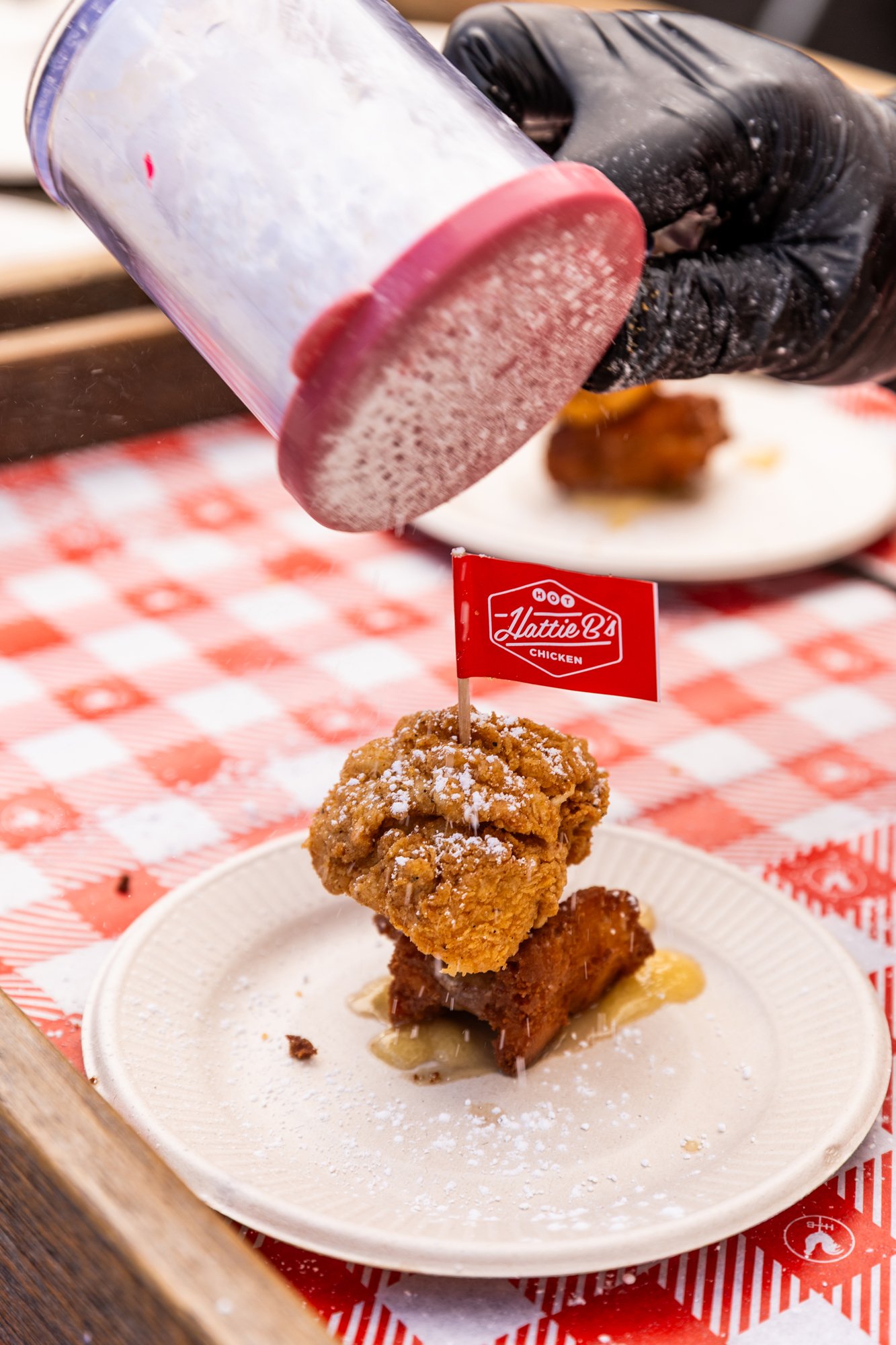 A piece of fried chicken and a piece of cooked pork belly on a plate, garnished with powdered sugar and a small flag labeled 'Hattie B's Hot Chicken' with a red and white checkered tablecloth underneath. Southbound Food Festival in Birmingham, Alabam