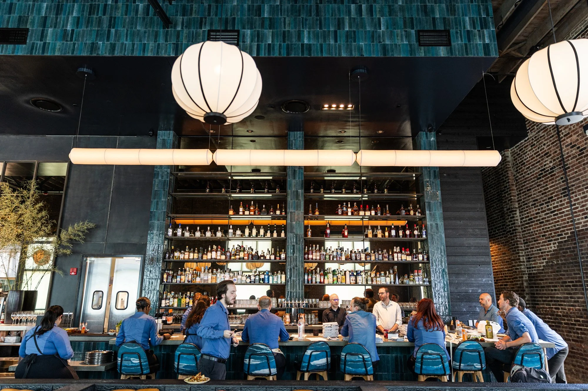 People sitting and standing at a bar in a restaurant with a large bottle display shelf behind the bar, dark brick walls, and modern hanging light fixtures.