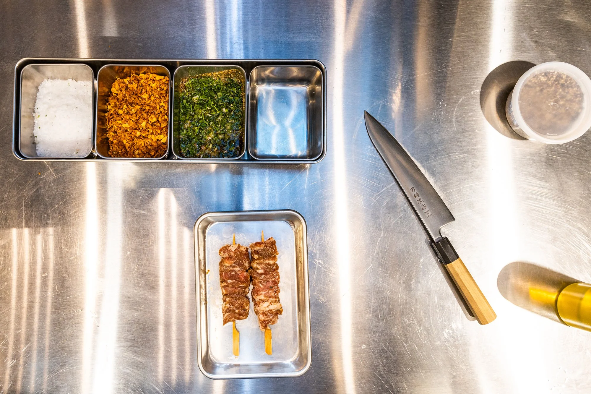 A stainless steel countertop with a metal tray containing salt, crushed garlic, chopped green herbs, and an empty compartment. A small plate with two skewered pieces of grilled meat, a kitchen knife, a plastic cup with seasonings, and a yellow squeez