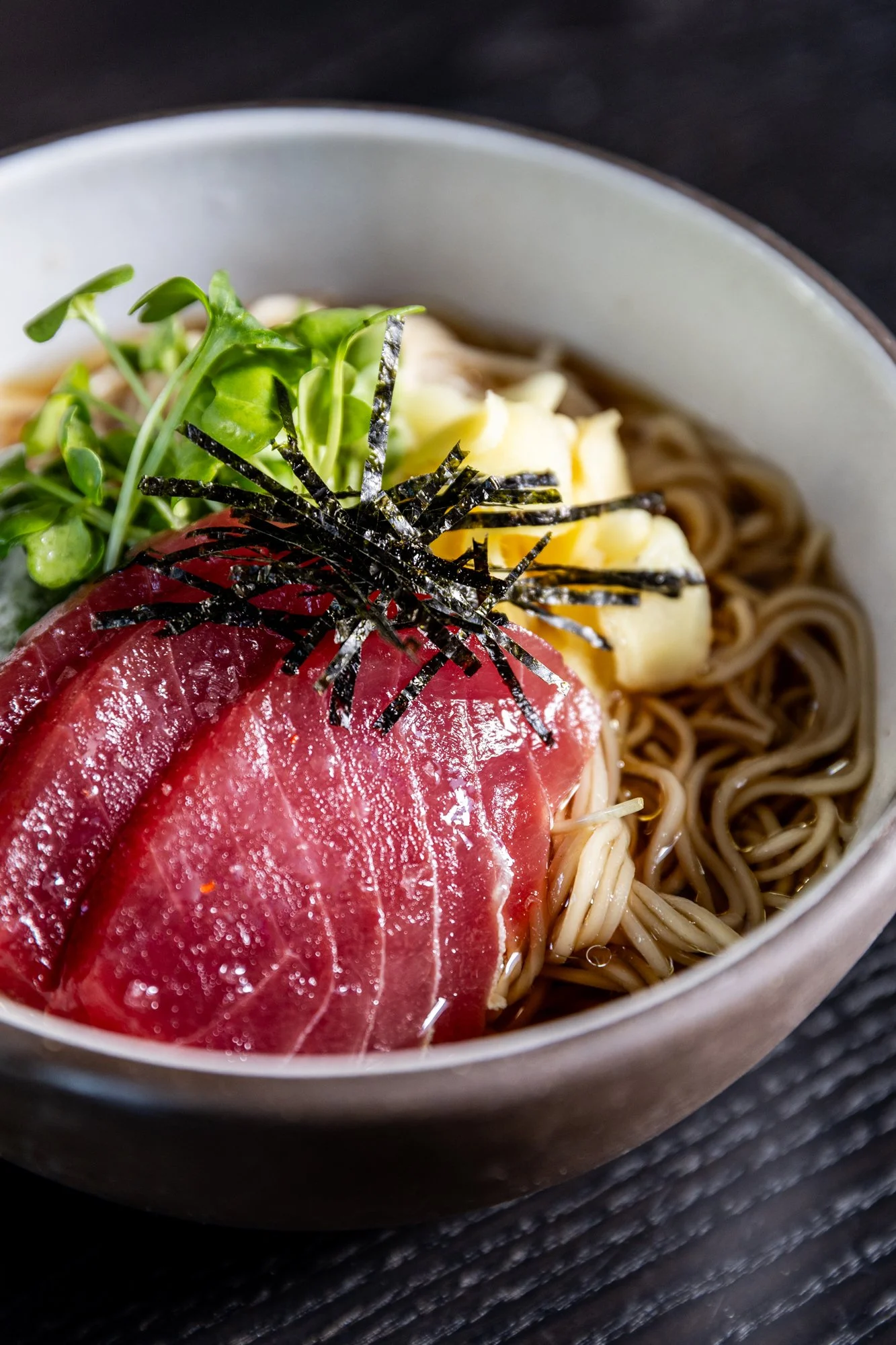 Bowl of Japanese soba noodles topped with slices of raw tuna, shredded nori, scrambled eggs, and microgreens.