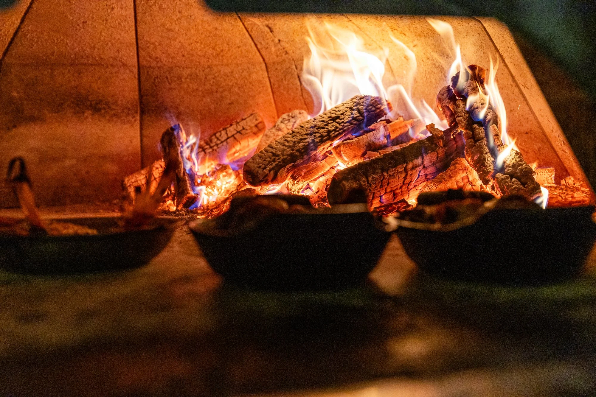 A close-up of burning wood logs with visible flames in a fire pit or fireplace. Ovenbird Restaurant in Birmingham, Alabama.