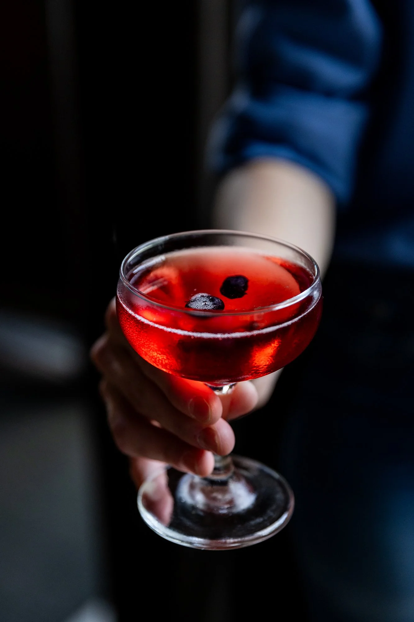 A person holding a cocktail glass filled with a red drink, garnished with blueberries, in a dimly lit setting. Hot and Hot Fish Club Restaurant in Birmingham, Alabama.