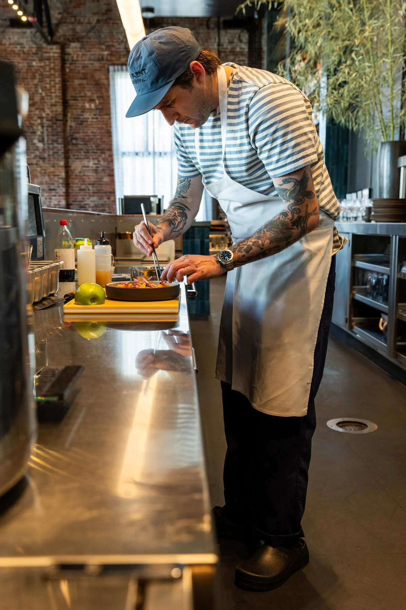 A male chef with tattoos, wearing a gray cap, striped t-shirt, white apron, and black shoes, preparing food at a stainless steel counter in a restaurant kitchen with brick walls, plants, and large windows.