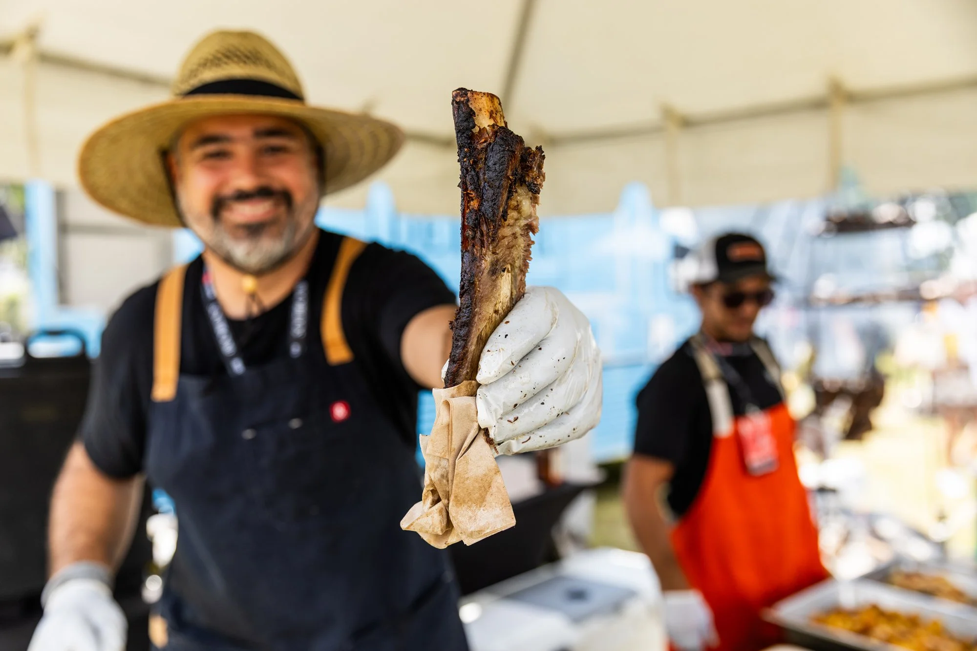 Man holding a large, charred piece of meat at a food stand, smiling at the camera, with another person in the background wearing sunglasses and an apron. Southbound Food Festival in Birmingham, Alabama.