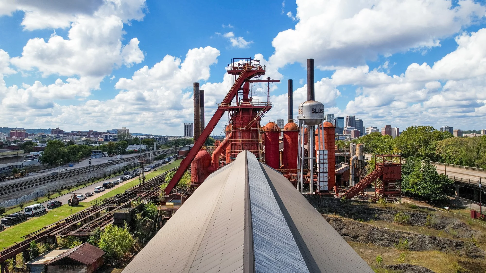 A bird's eye view of an industrial factory with red metal tanks, pipes, and machinery, with a modern city skyline in the background under a partly cloudy sky. Southbound Food Festival in Birmingham, Alabama.