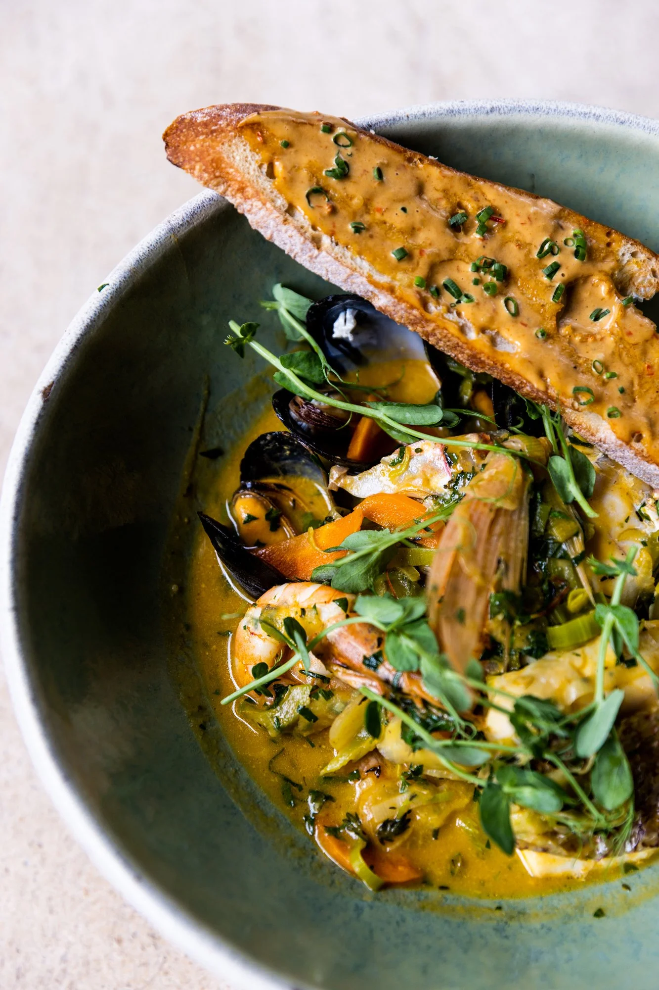 A bowl of seafood soup with shrimp, mussels, and vegetables garnished with herbs, accompanied by a slice of toasted bread with cheese and herbs. Hot and Hot Fish Club Restaurant in Birmingham, Alabama.