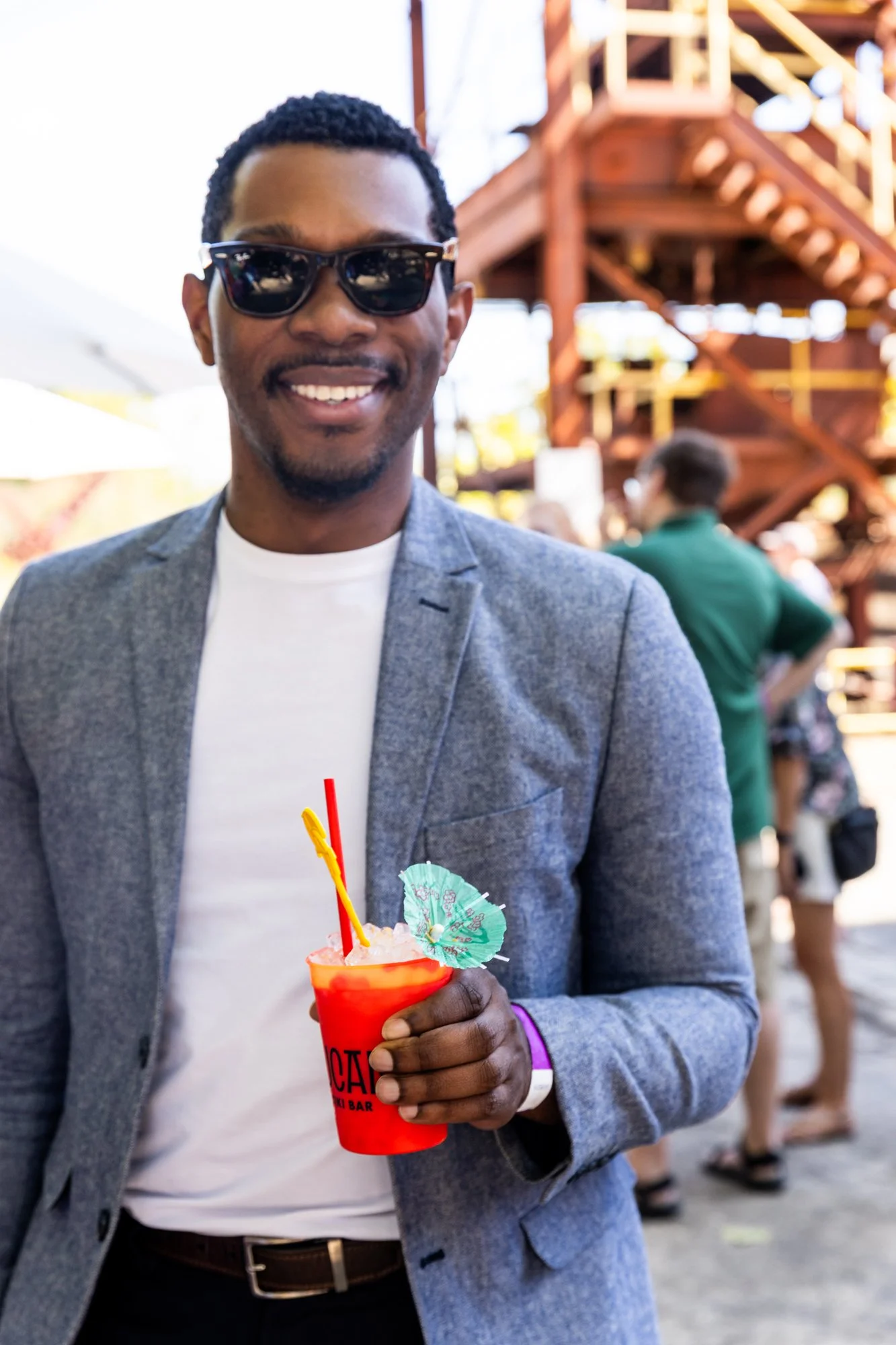 A smiling man wearing sunglasses and a grey blazer holding a red tropical cocktail with a mini umbrella and straws at an outdoor event. Southbound Food Festival in Birmingham, Alabama.