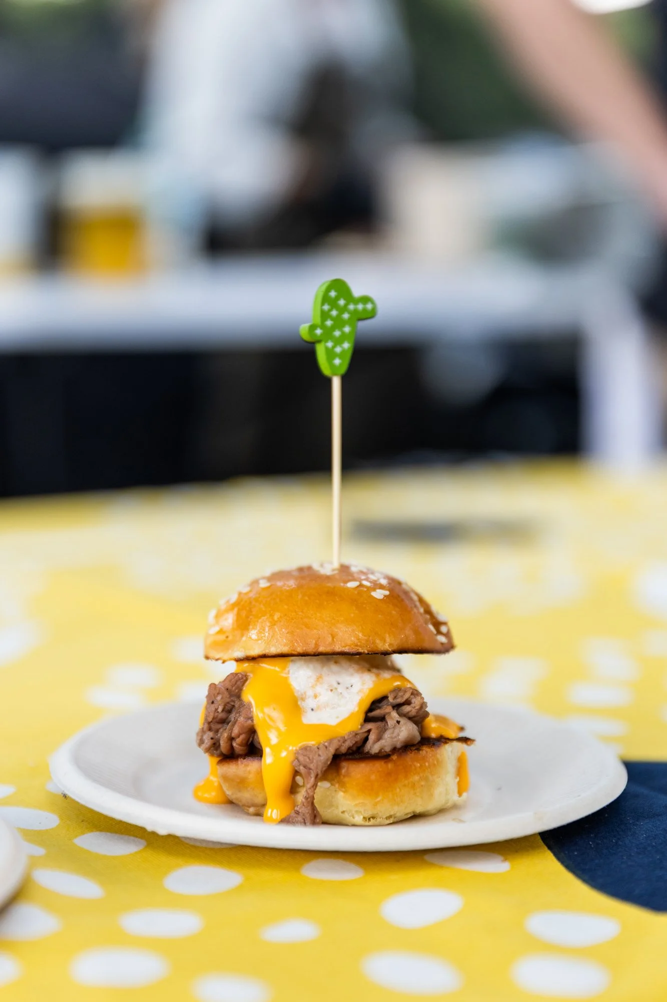 Mini cheeseburger slider with melted cheese, a beef patty, and a fried egg on a white plate, garnished with a green cactus-shaped toothpick, on a yellow polka dot tablecloth. Southbound Food Festival in Birmingham, Alabama.