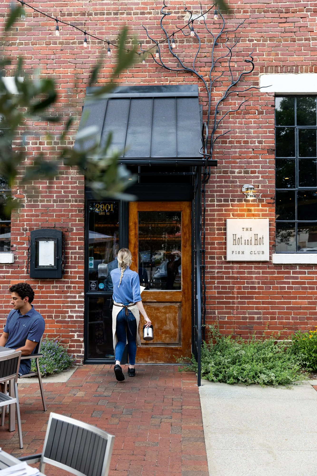 People entering and sitting outside a restaurant called 'The Hot and Hot Fish Club,' located in a brick building with outdoor string lights and a decorative metal tree on the wall. Hot andHot Fish Club Restaurant in Birmingham, Alabama.