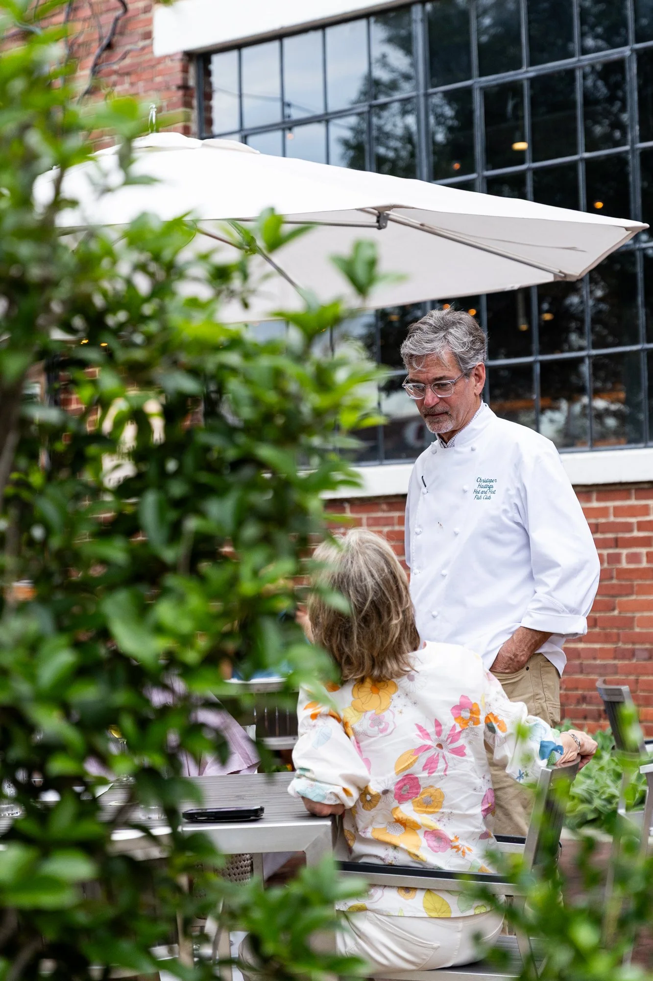A chef talking to a woman sitting at a table outside at a restaurant, with greenery in the foreground and a brick building with large windows in the background Hot andHot Fish Club Restaurant in Birmingham, Alabama.