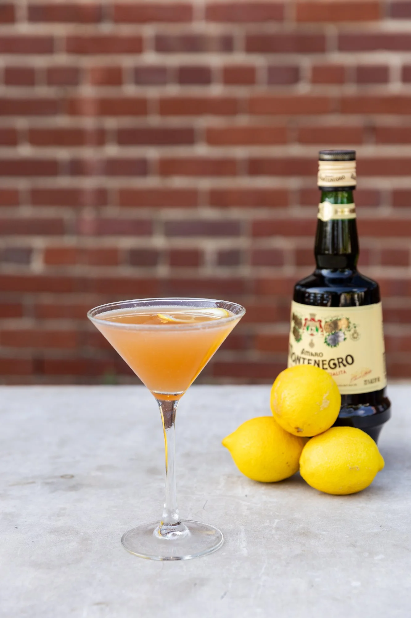 A cocktail glass with a citrus drink and lemon slices, set on a white surface with three lemons and a bottle of Amaro Montenegro in the background, against a brick wall. Hot and Hot Fish Club Restaurant in Birmingham, Alabama.