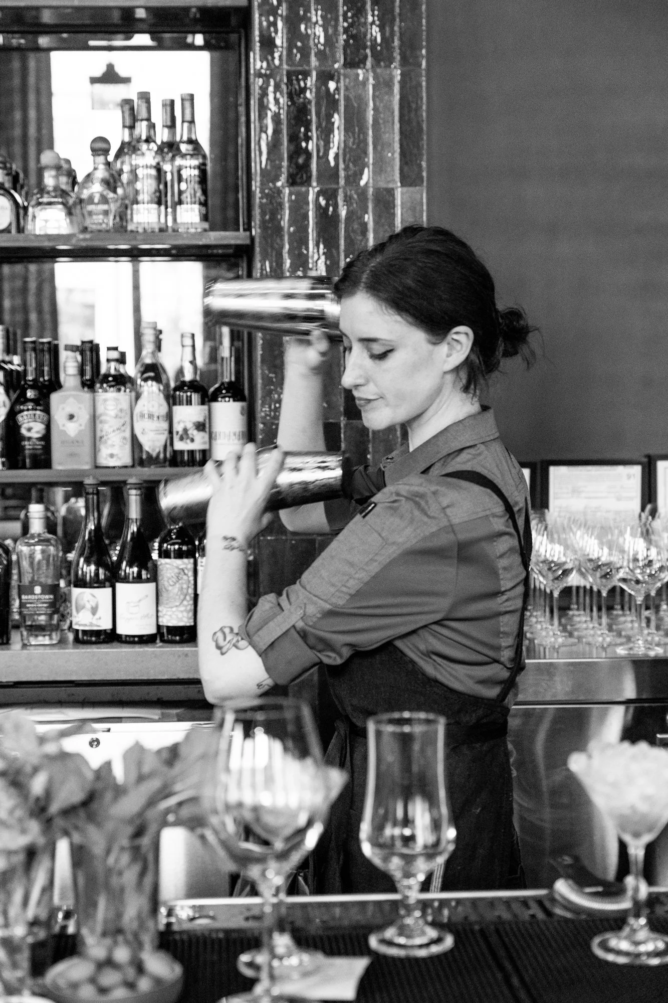 A female bartender with dark hair tied back, wearing a rolled-up long sleeve shirt, is pouring a drink into a cocktail shaker behind a bar filled with wine bottles, glasses, and garnishes in a restaurant or bar setting.