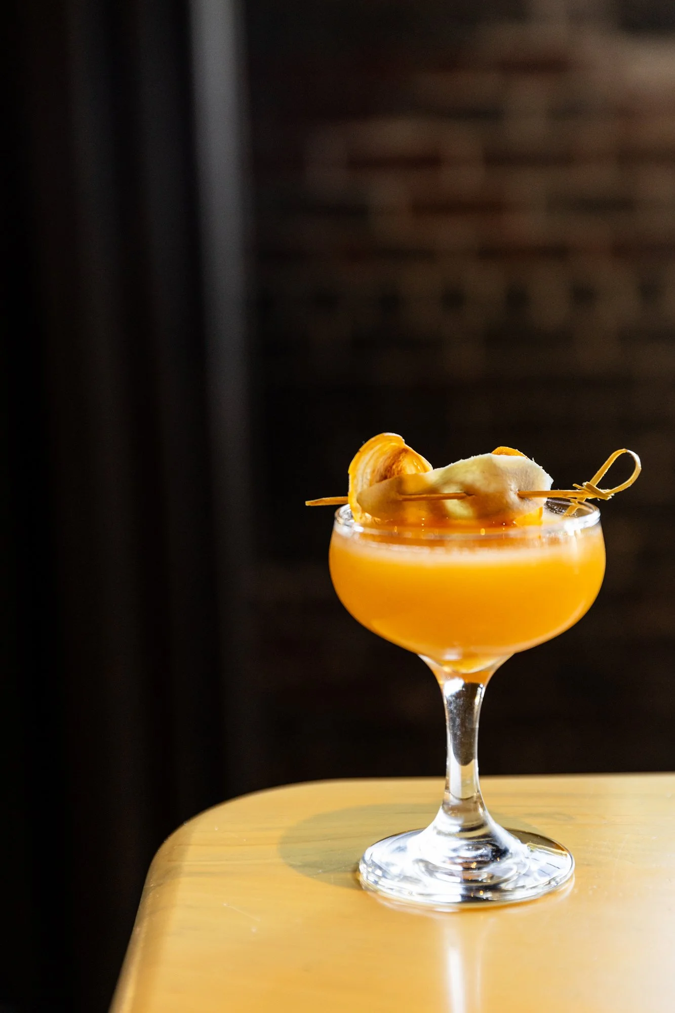 A yellow-orange cocktail in a coupe glass garnished with a slice of banana and a cocktail skewer, placed on a wooden surface. Hot and Hot Fish Club Restaurant in Birmingham, Alabama.
