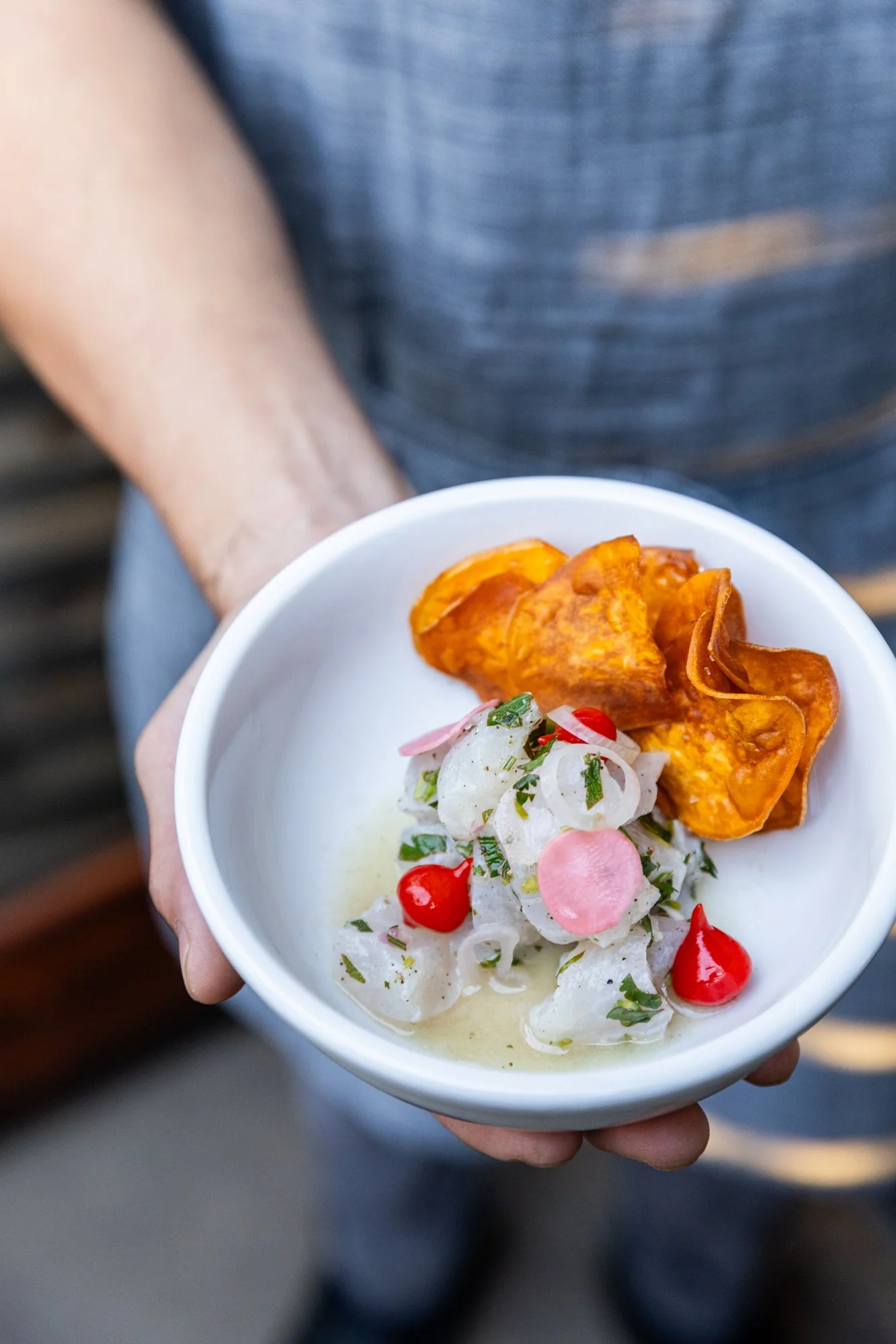 Person holding a bowl of ceviche topped with sliced onions, cilantro, red chili, and served with sweet potato chips. Ovenbird Restaurant in Birmingham, Alabama.