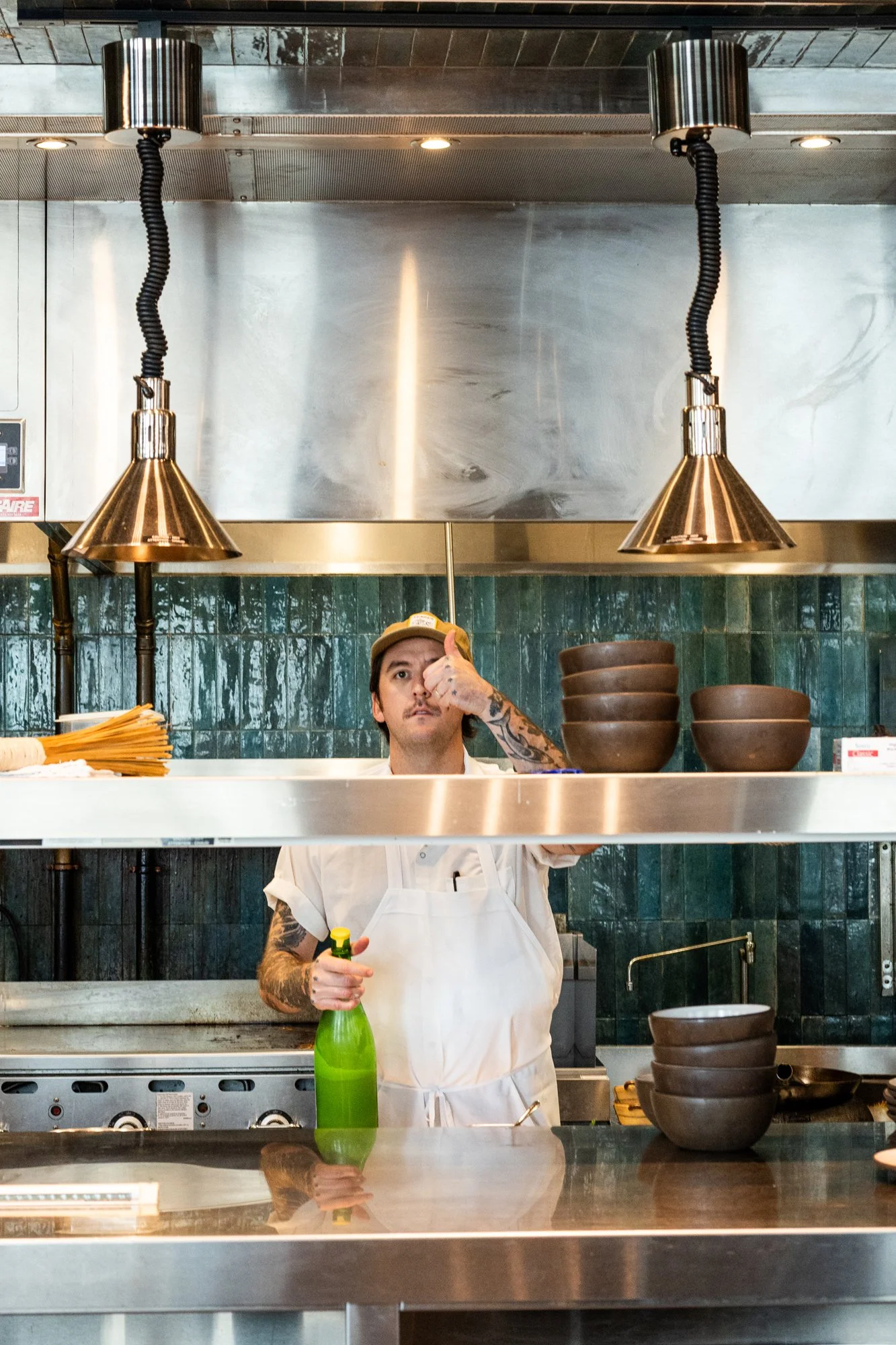 A chef with tattoos giving a thumbs up behind a kitchen counter with stacked bowls and cooking utensils.